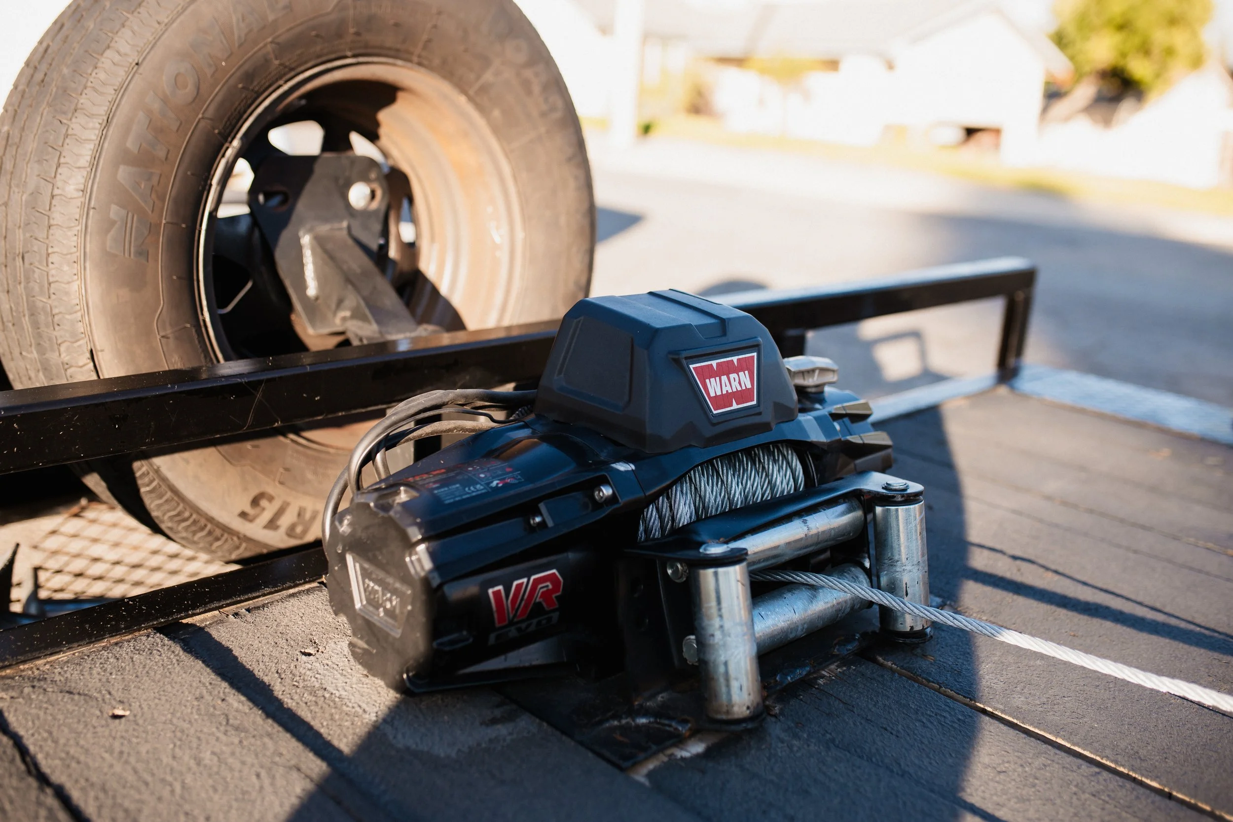 Close-up of a trailer winch with a cable, mounted on a flatbed trailer, with a large tire in the background.