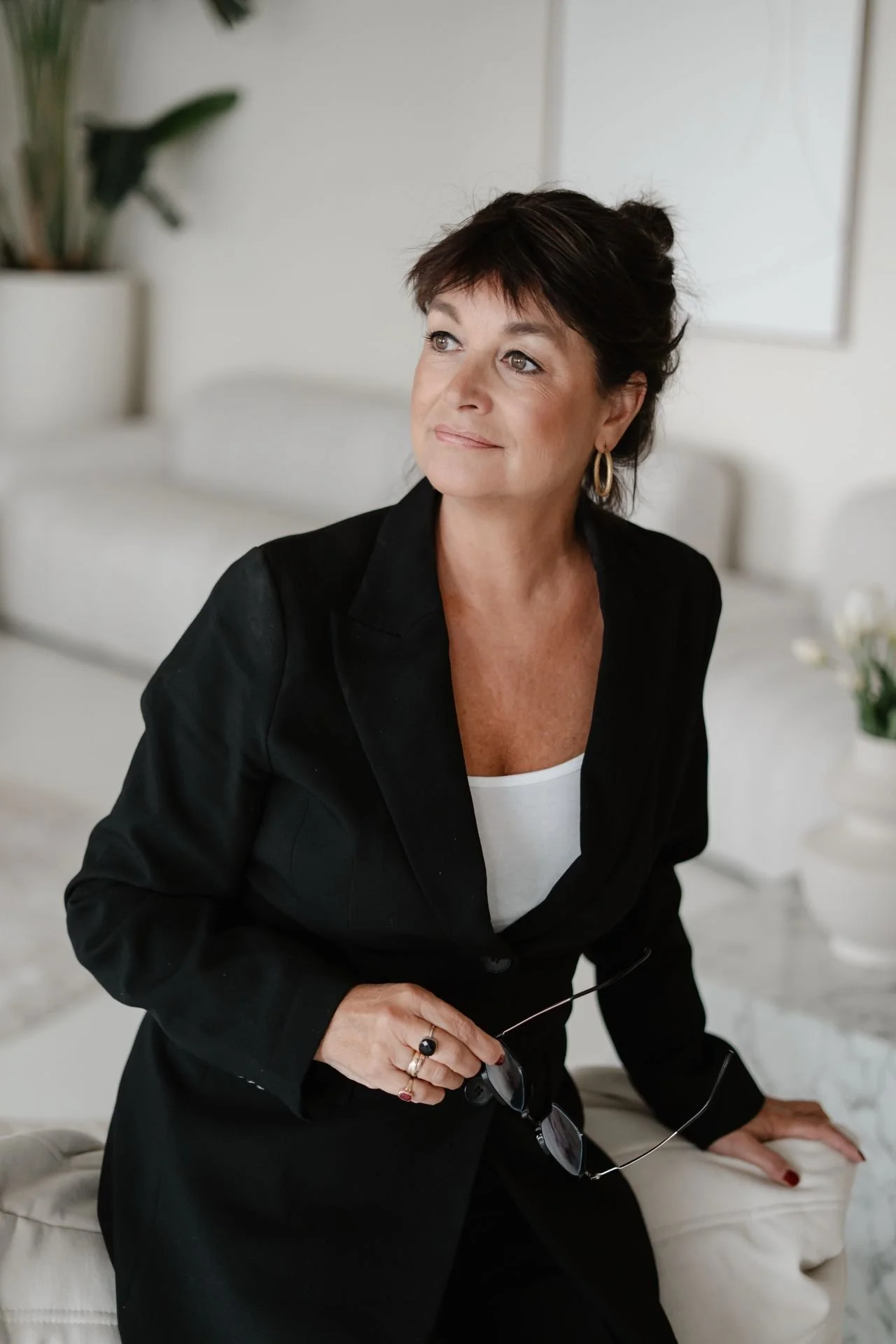 Portret van een vrouw in een lichte, serene ruimte, gekleed in een zwart jasje en witte top, met een bedachtzame blik opzij en een kalme, zelfverzekerde uitstraling.