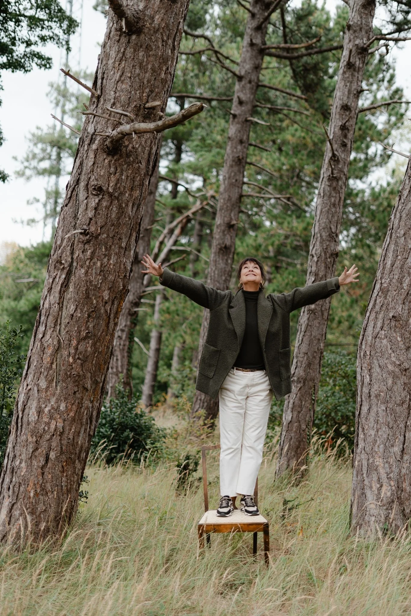 Vrouw in een groen jasje en lichte broek staat buiten op een houten stoel tussen hoge bomen. Ze spreidt haar armen en kijkt omhoog, omringd door gras en een rustig, natuurlijk boslandschap.