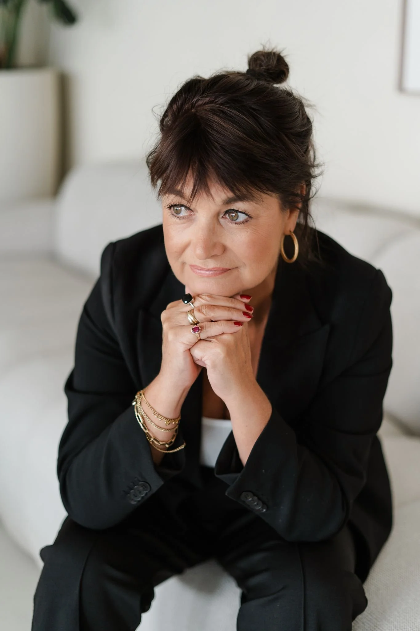 Close-up portret van een vrouw in een zwart pak, zittend en voorover geleund met haar handen onder haar kin, terwijl ze bedachtzaam opzij kijkt in een lichte, rustige ruimte.