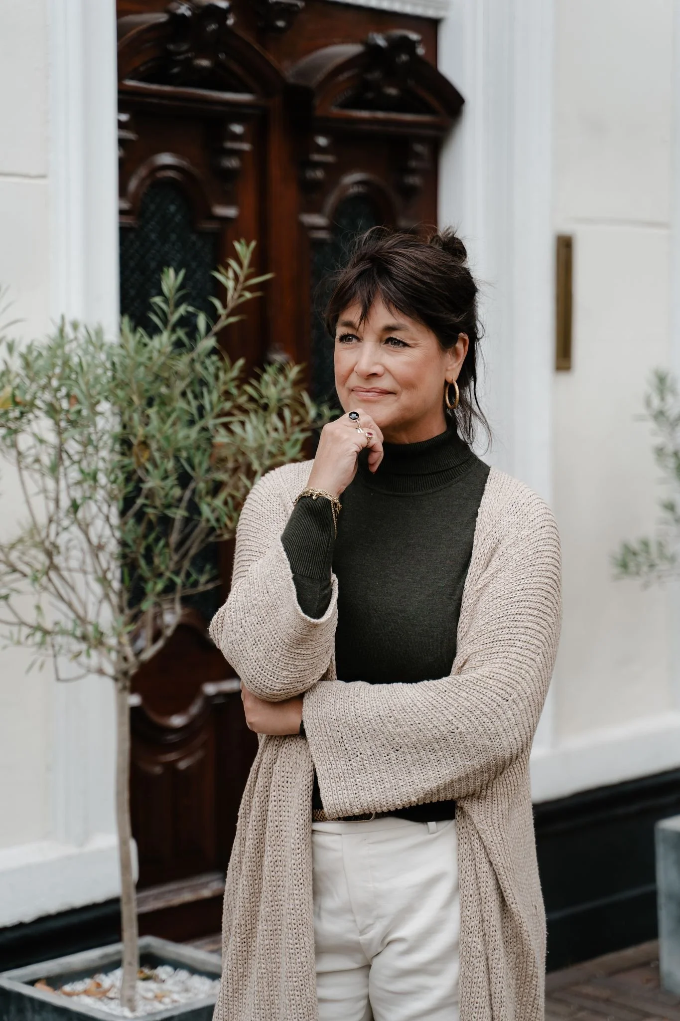 Vrouw staat voor een klassieke houten deur en kijkt bedachtzaam voor zich uit. Ze draagt een donkergroene coltrui met een lichte, gebreide vest en een lichte broek, met haar hand rustend onder haar kin en haar armen losjes gekruist.