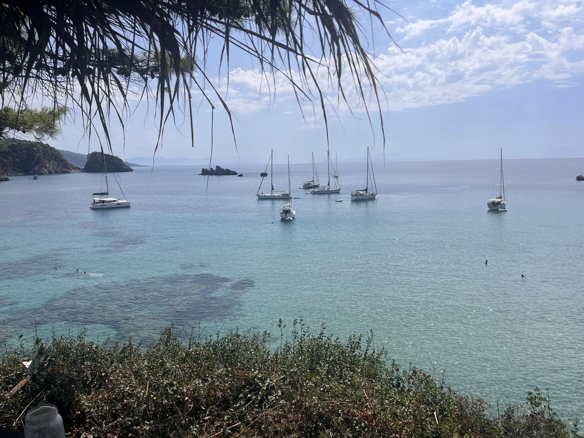 Rustige baai met helderblauw water, meerdere zeilboten voor anker en een rotsachtige kust op de achtergrond.