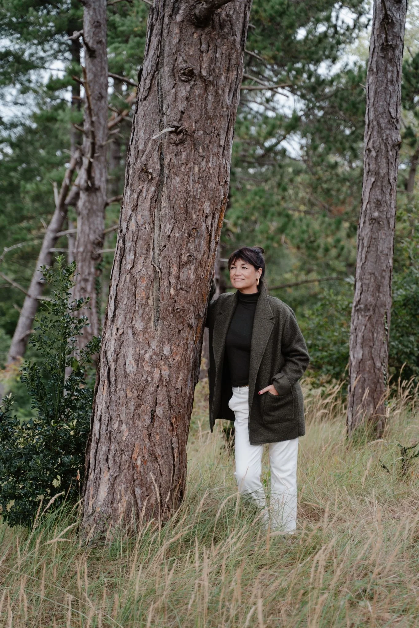 Vrouw in een groen jasje en lichte broek staat in een bos naast een hoge boom. Ze leunt licht tegen de boomstam, heeft één hand in haar zak en kijkt rustig voor zich uit tussen het gras en de bomen.