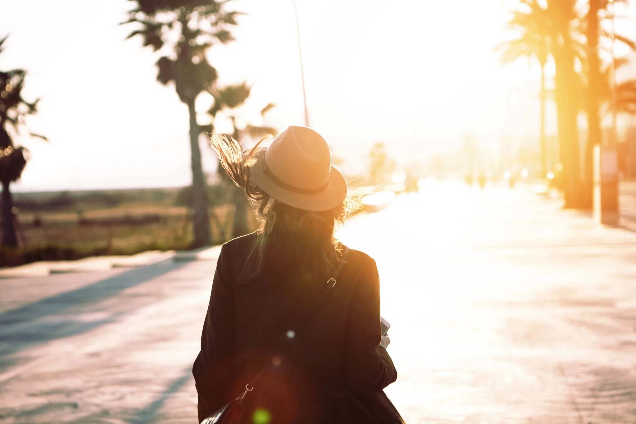 Persoon met hoed loopt in tegenlicht over een zonnige boulevard, met palmbomen en een warme gloed van de ondergaande zon.