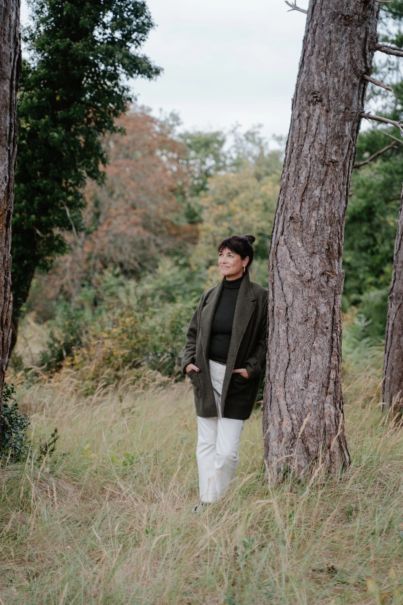 Vrouw staat ontspannen in een open bosrijke omgeving tussen hoge bomen en gras. Ze draagt een donkergroene coltrui met een bijpassende jas en lichte broek, met haar handen in haar zakken, terwijl ze rustig de verte inkijkt.