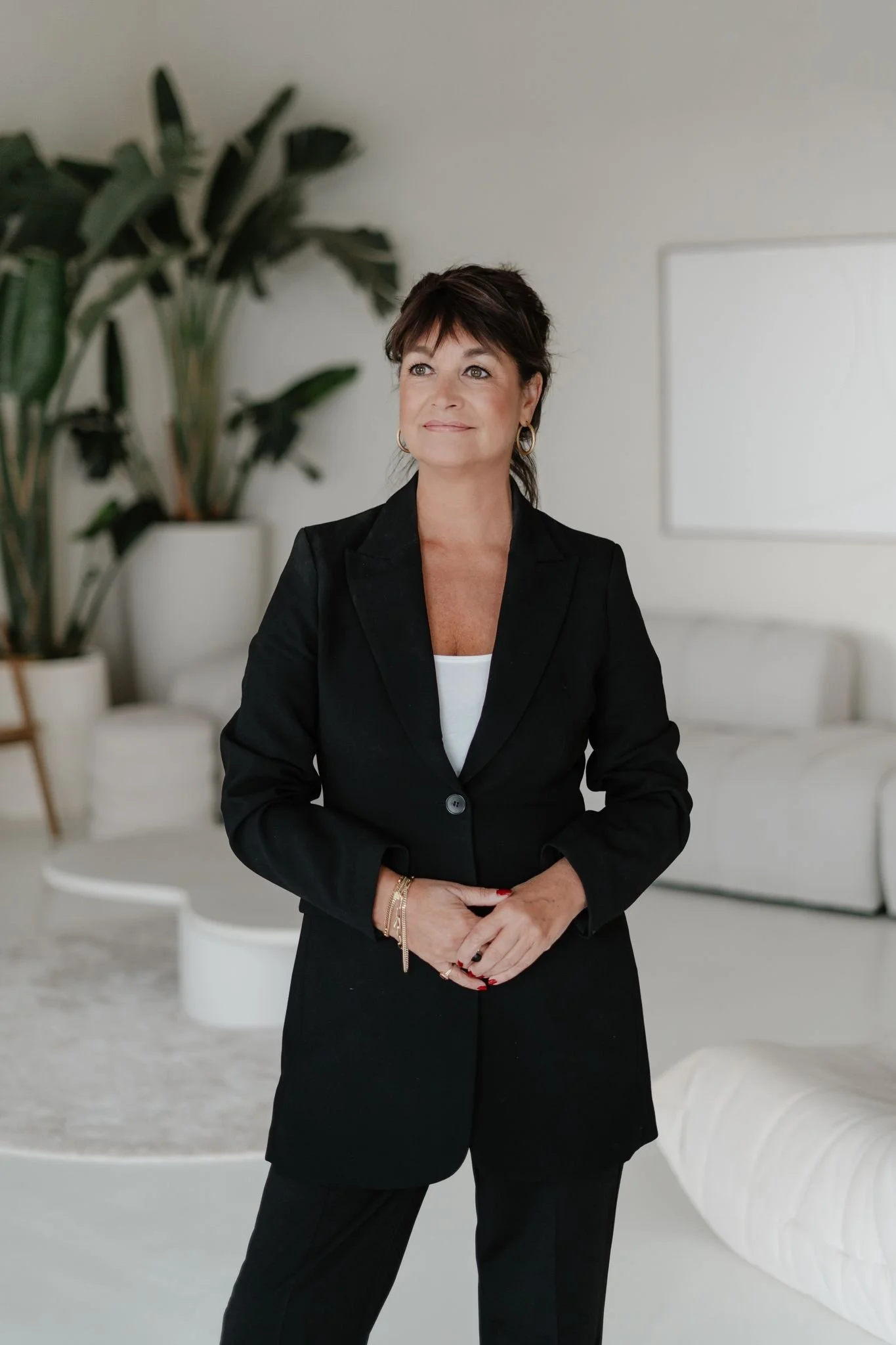 Vrouw in een elegant zwart pak met witte top staat ontspannen in een lichte, minimalistische ruimte met zachte witte meubels en grote groene planten. Ze kijkt rustig iets omhoog en straalt zelfvertrouwen en kalmte uit in een moderne, serene setting.