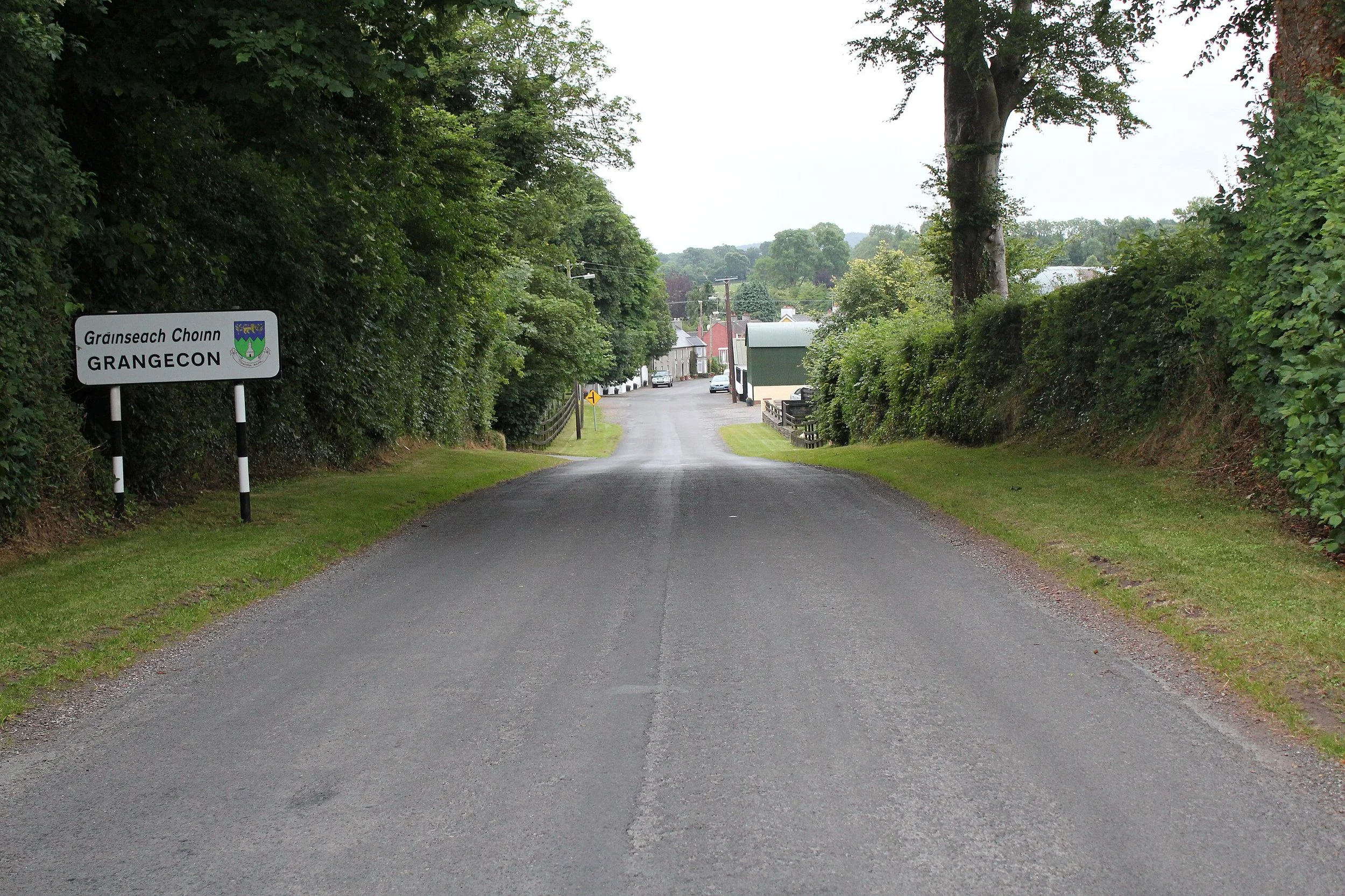 A rural road with a village sign on the left that reads 'Gráinseach Chonn' and 'GRANGECON' in black letters on a white background, surrounded by green trees and bushes, leading to a small town with buildings and cars in the background.