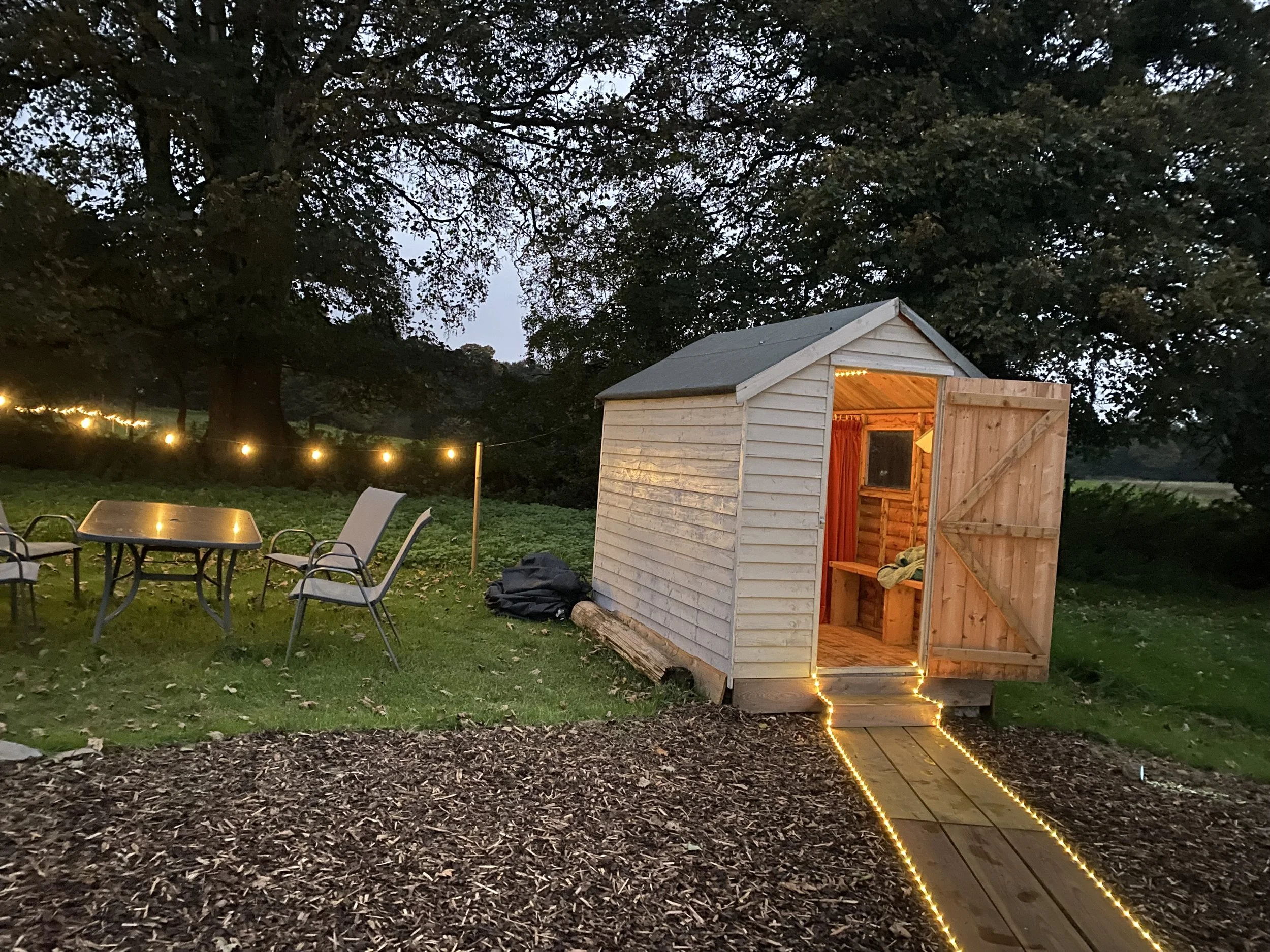 Hot Rocks Sauna wooden shed with an open door illuminated by warm yellow lights, set outdoors at dusk.