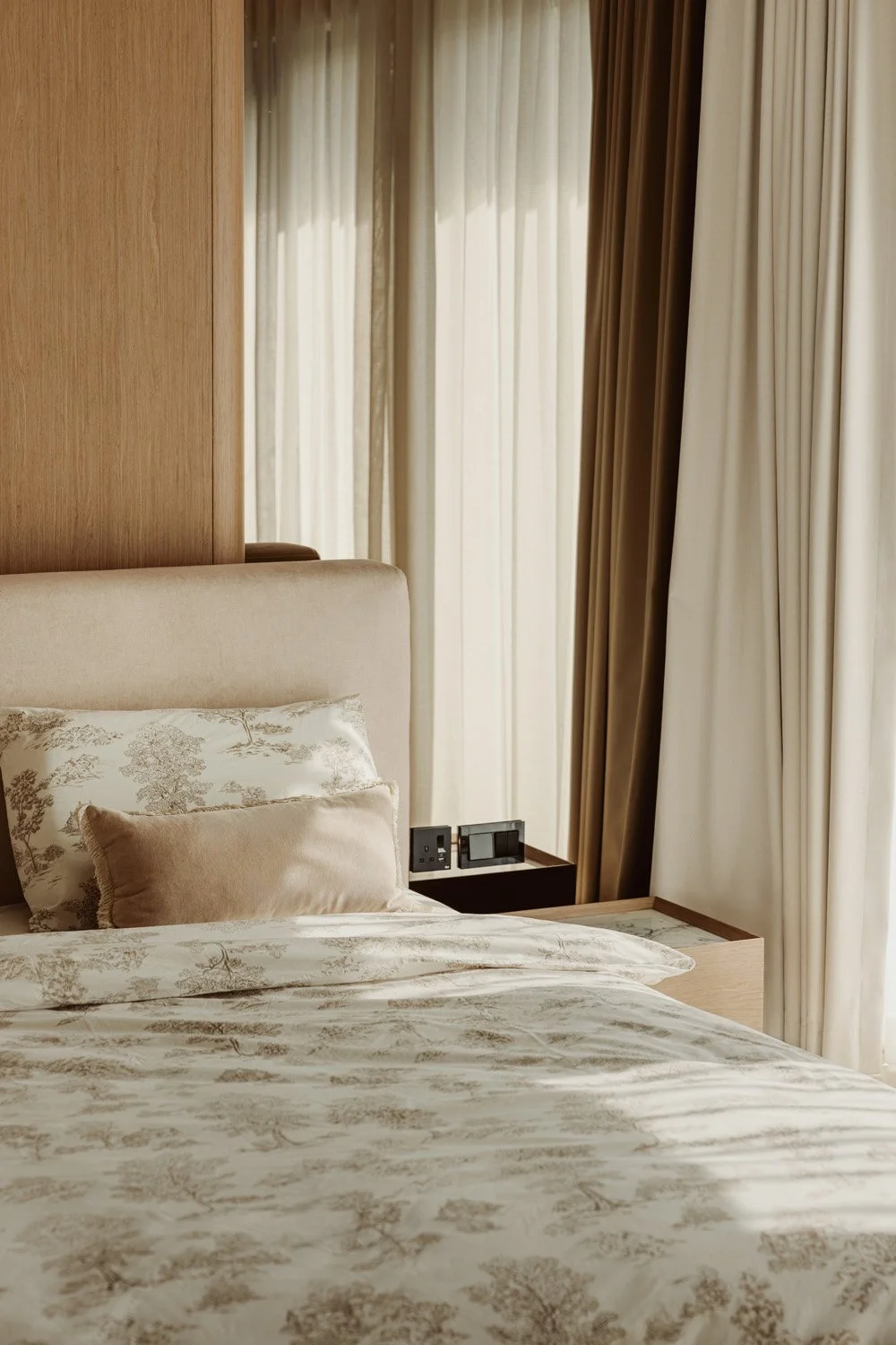 Vignette detail of a luxury master bedroom with toile de Jouy patterned bedding, a velvet accent cushion, oak veneer headwall, and layered sheer and blackout curtains, designed by Limestone in Dubai
