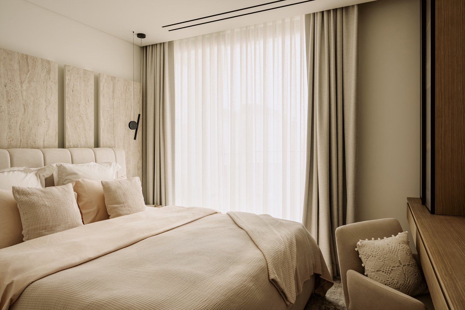 Luxury guest bedroom with travertine panel feature wall, upholstered bouclé headboard, warm neutral bedding, and sheer floor-to-ceiling curtains by Limestone