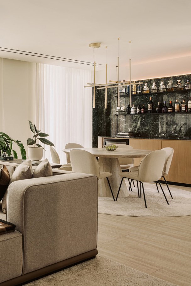 Luxury open-plan living and dining room designed by Limestone Dubai, featuring a travertine round dining table, upholstered chairs, a green marble home bar, and a sculptural brass chandelier