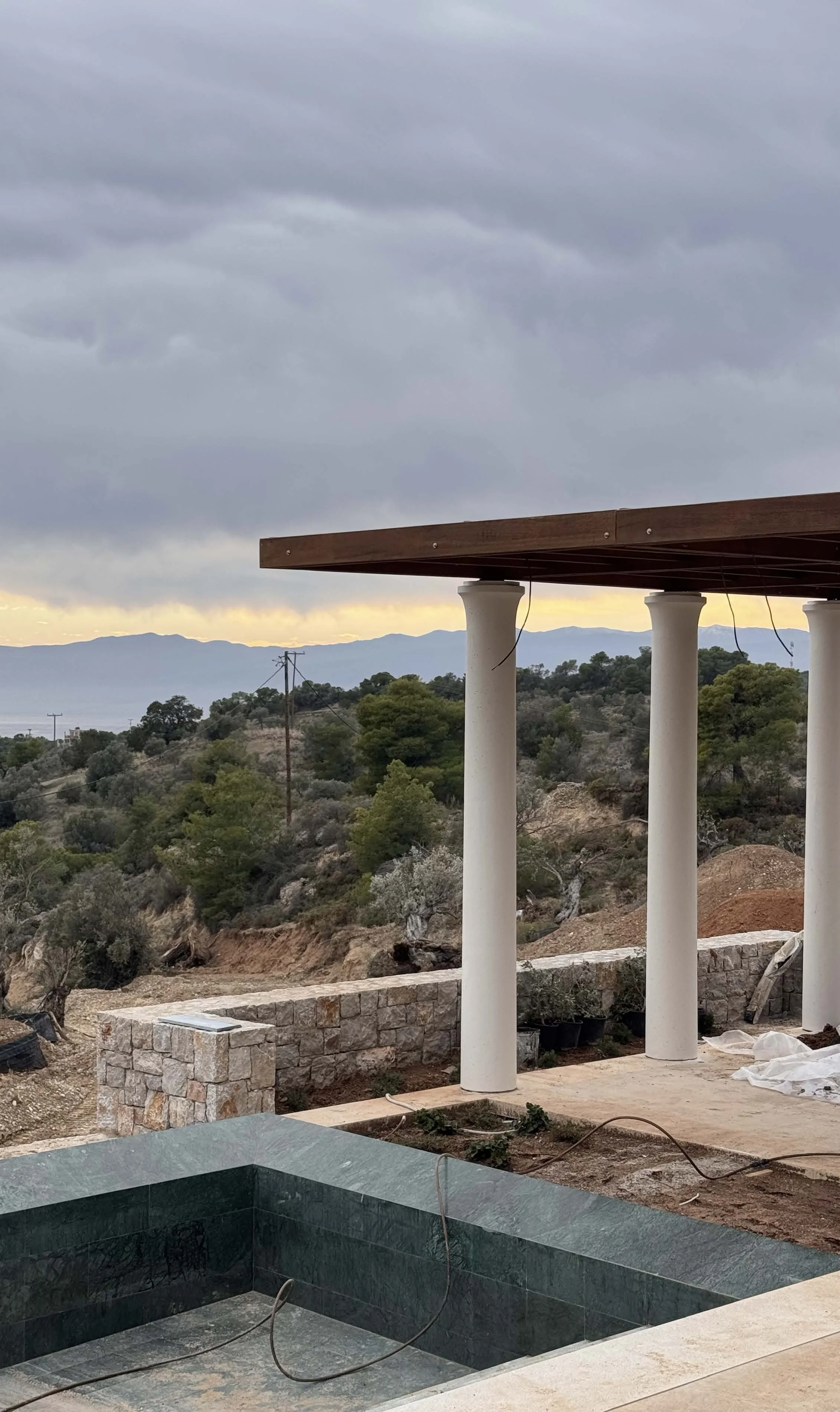 Construction site with a stone wall, white columns supporting a wooden roof, and an empty pool in the foreground, with a landscape of trees, power poles, and mountains under a cloudy sky in the background.