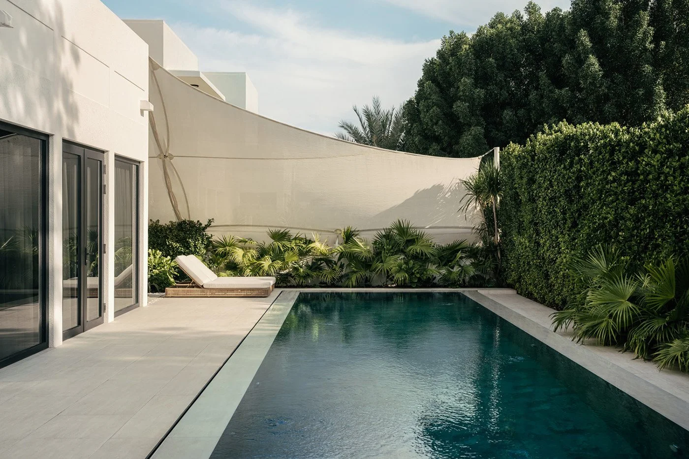 Wide exterior view of a luxury villa lap pool with a sculptural sail shade, dense tropical hedge screening, palm trees, and a sun lounger, designed by Limestone in Dubai