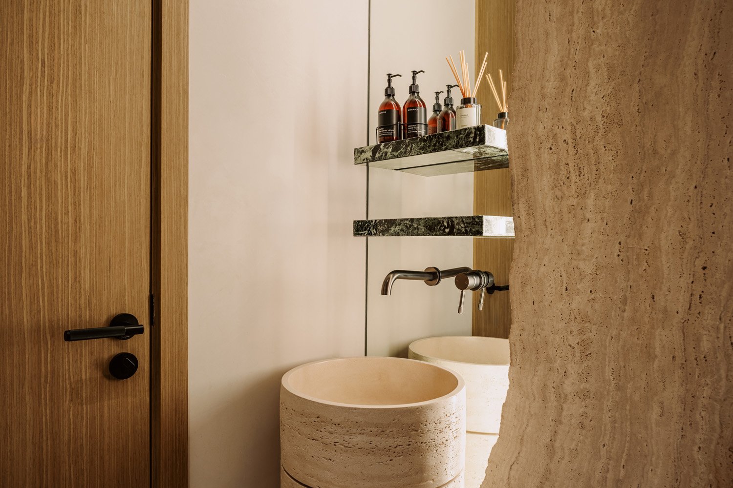 Luxury powder room featuring a sculptural travertine vessel basin, verde marble floating shelf, wall-mounted brushed steel tapware, and oak door by Limestone
