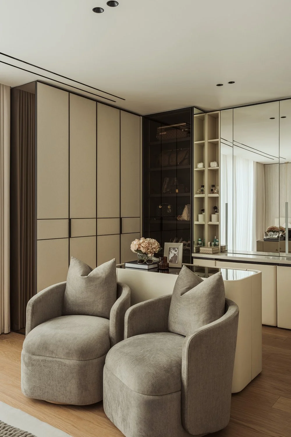 Her dressing room in Villa Ambra, Dubai, with fabric-wrapped wardrobes outlined in dark metal, a glass display cabinet, a leather-wrapped island and two grey armchairs.