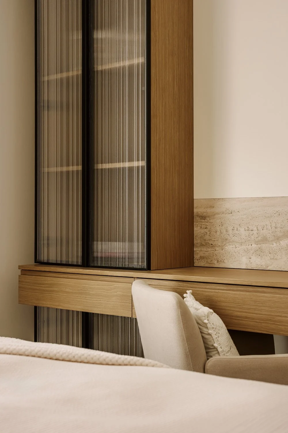 Close-up detail of bespoke bedroom joinery featuring reeded glass wardrobe doors, oak timber frame, floating desk, and travertine headwall by Limestone