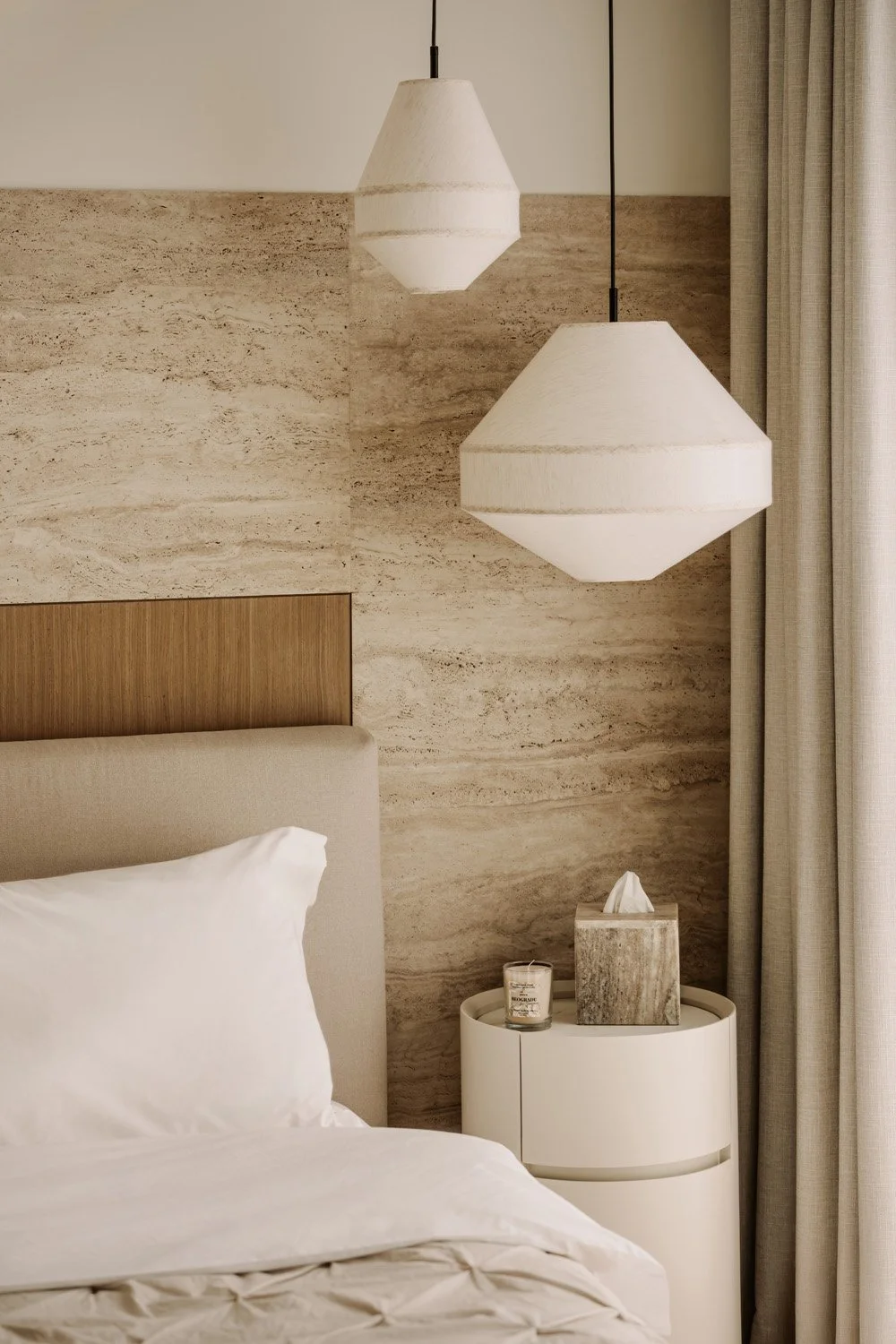 Detail of a luxury bedroom with travertine stone headwall, sculptural white pendant lights, and a cylindrical bedside table by Limestone