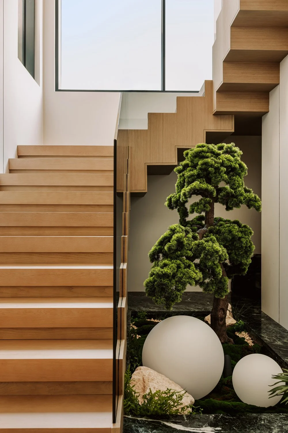 Architectural staircase void featuring cantilevered oak treads, a sculptural bonsai tree planted in a green marble base with globe lights, designed by Limestone