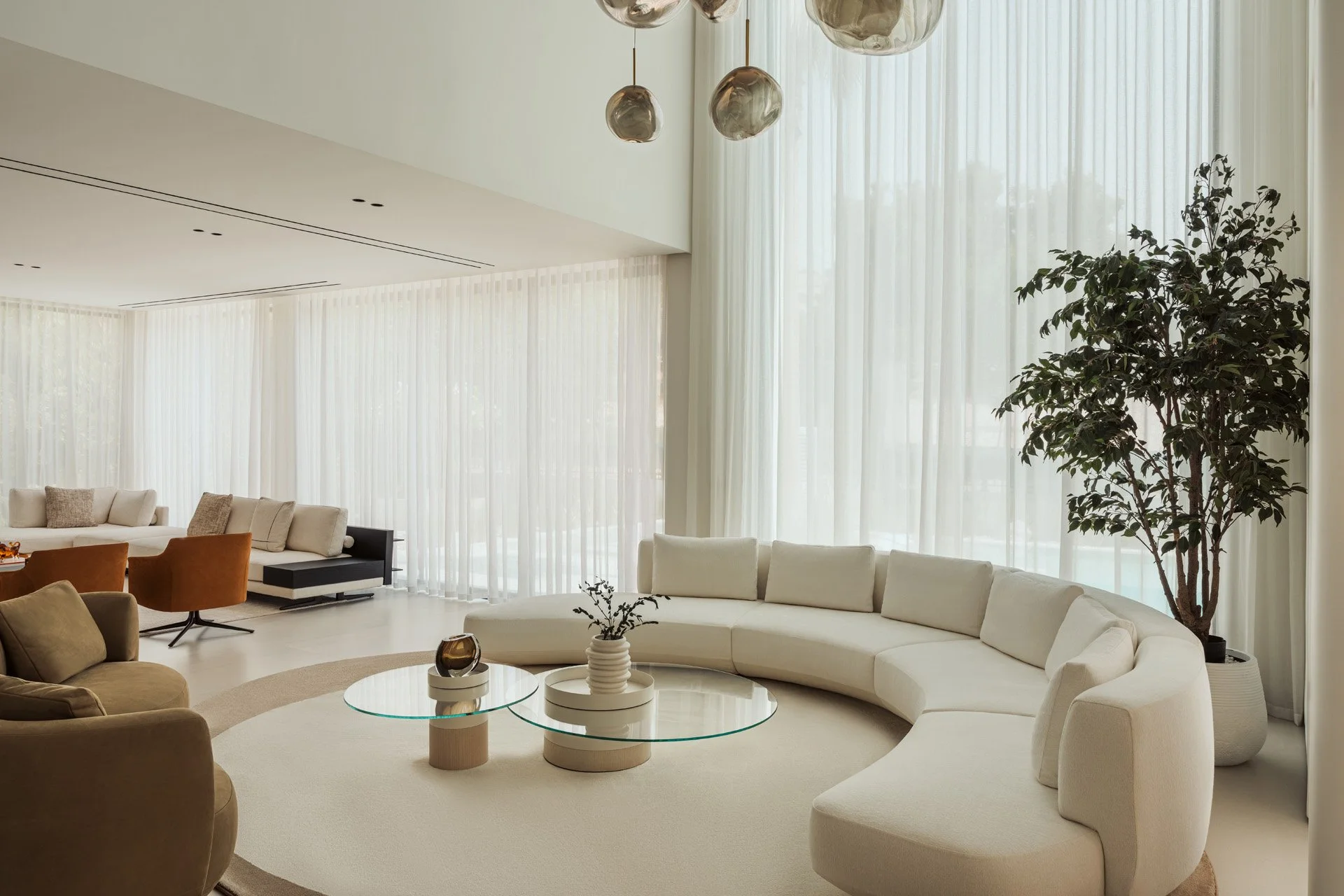 Open-plan living space in Villa Ambra, Jumeirah Islands, Dubai: a double-height lounge with a curved cream sofa, glass coffee tables and smoked glass pendants, opening onto a second seating area.