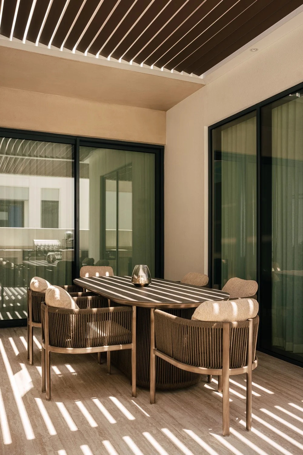 Luxury covered outdoor dining terrace with a louvred timber pergola, oval stone-top table, and woven rope dining chairs casting striped shadows, designed by Limestone in Dubai