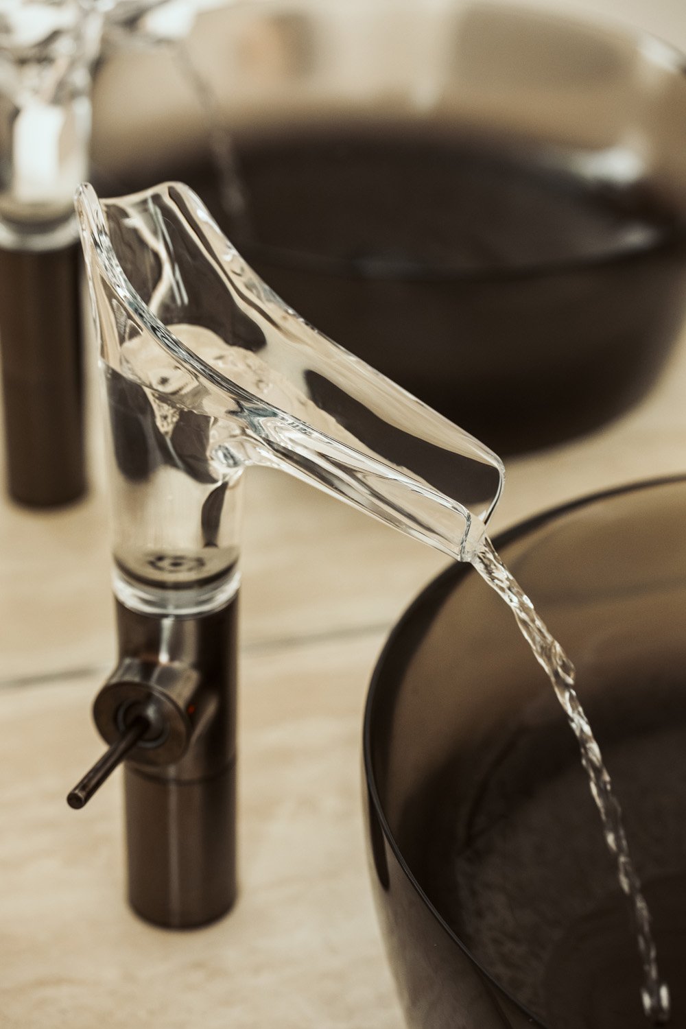 Close-up of a clear glass-spout tap pouring into a smoked-glass basin on a travertine vanity in Villa Ambra, Jumeirah Islands.