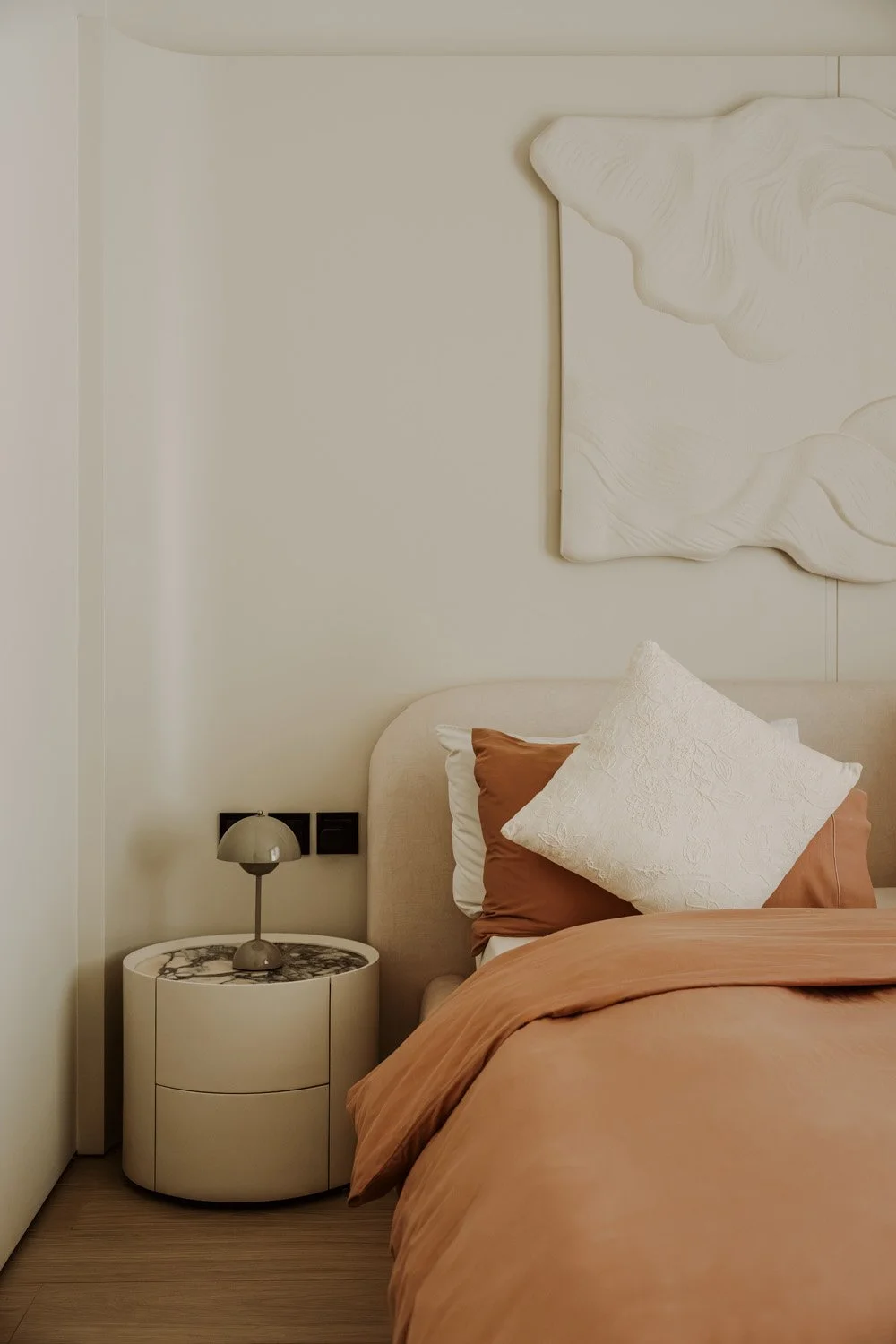 Bedside vignette of a luxury bedroom featuring a cylindrical marble-topped nightstand, a Flowerpot table lamp, terracotta silk pillowcases, and a white sculptural relief artwork, designed by Limestone in Dubai