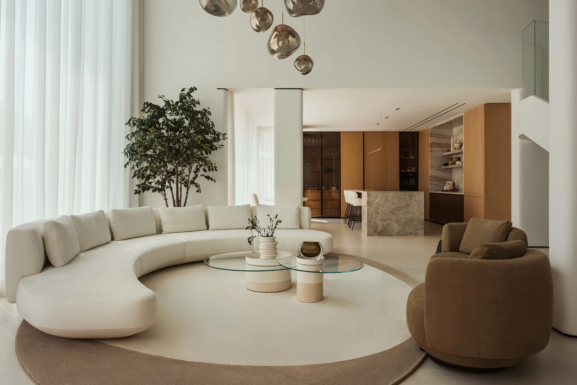 Double-height formal living room in Villa Ambra, Jumeirah Islands, with a curved cream sofa, glass coffee tables, a round rug and smoked glass pendants, opening onto the kitchen.