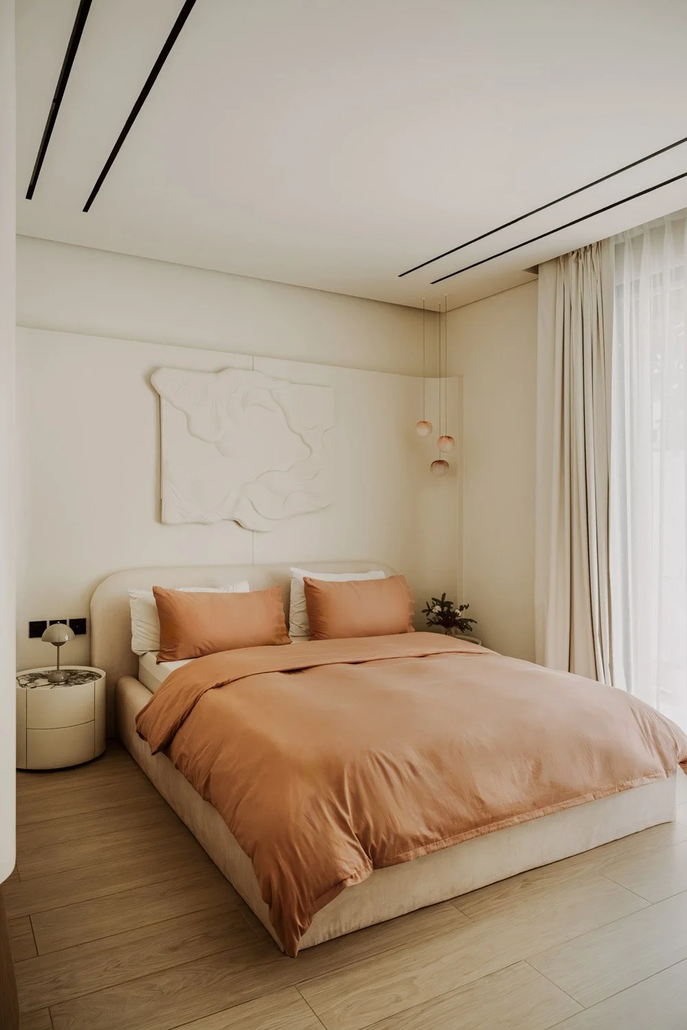 Full view of a luxury girl's bedroom with terracotta silk bedding, a curved bouclé headboard, white sculptural relief wall artwork, amber glass cluster pendants, and floor-to-ceiling sheer curtains, designed by Limestone in Dubai