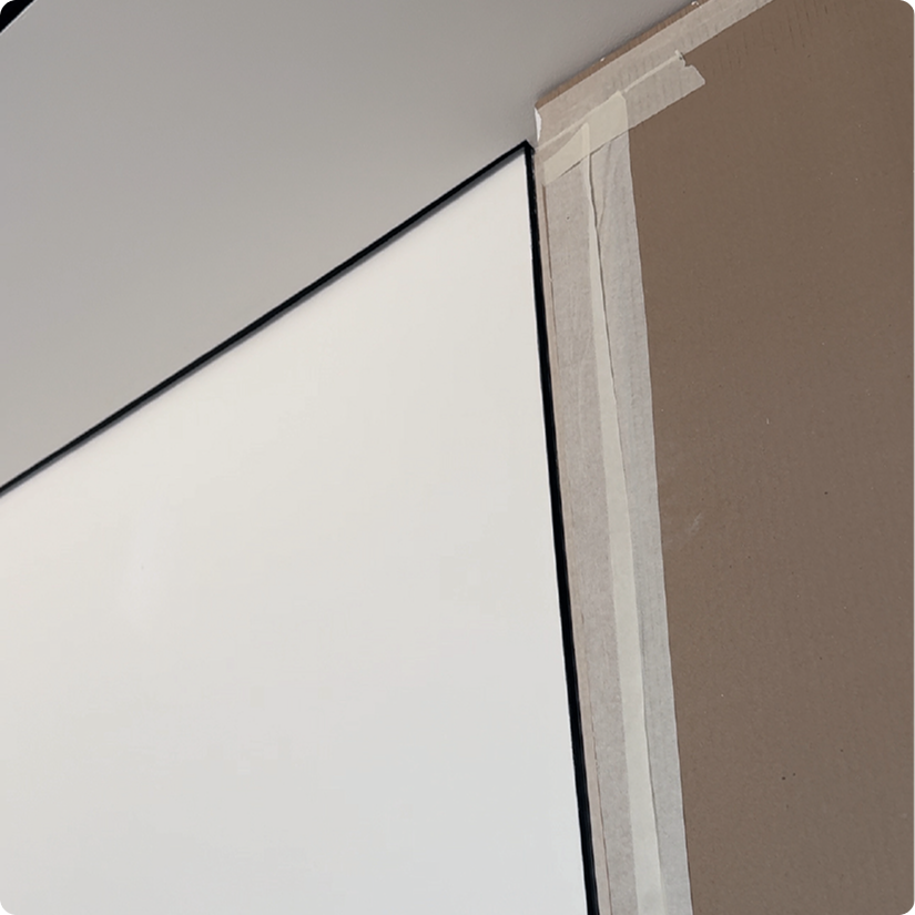 Close-up of a section of a room with a white wall, a portion of a ceiling, and a black-framed whiteboard or screen with painter's tape around the edges, suggesting it is being prepared for painting or installation.