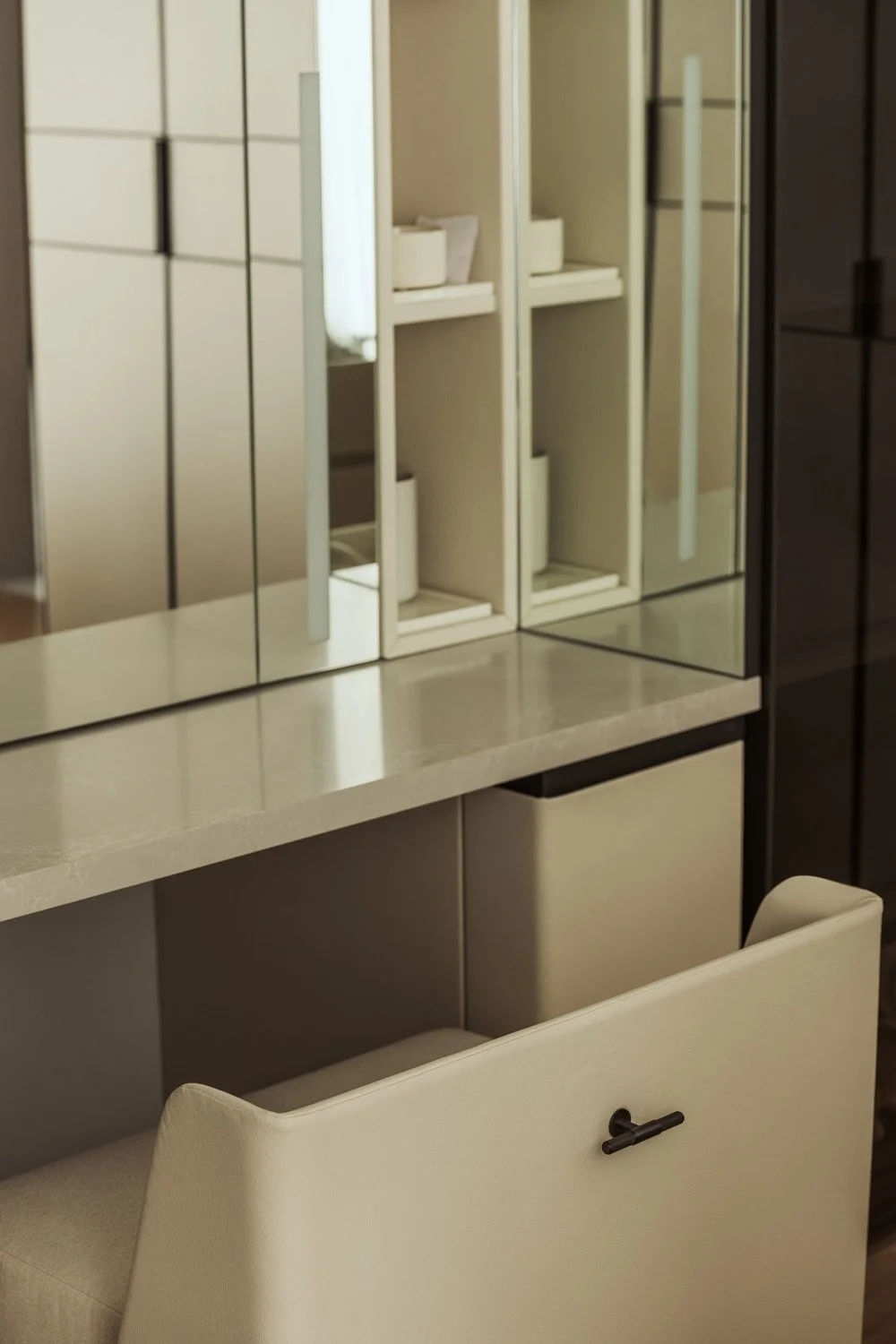 Dressing room vanity desk in Villa Ambra, Dubai, with a stone counter, mirrored panels with integrated lighting, open niche shelving and a leather pull-out chair with a dark metal handle.
