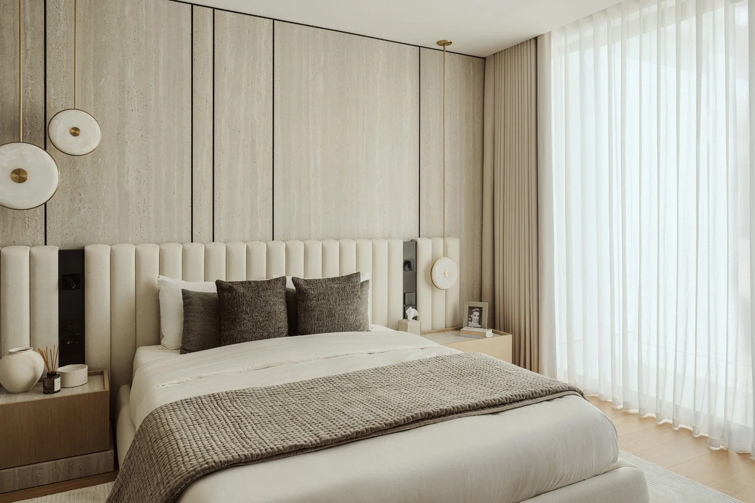 Master bedroom in Villa Ambra, Jumeirah Islands, with a travertine wall inlaid with dark metal lines, an upholstered channel headboard and alabaster disc pendants.