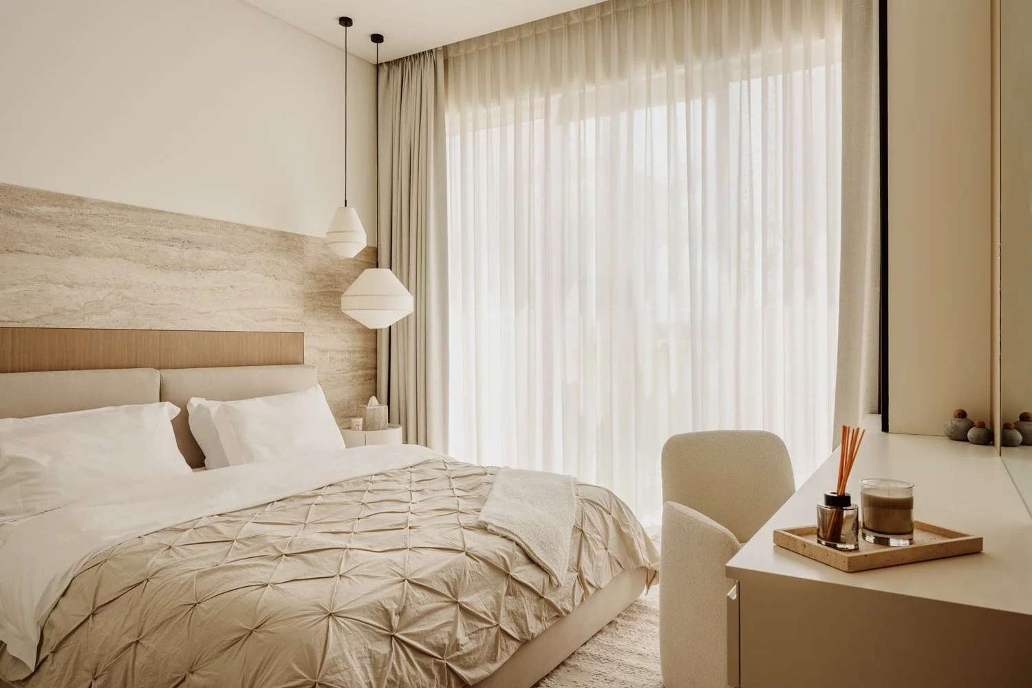 Full view of a luxury master bedroom with travertine stone feature wall, linen upholstered bed, pendant lights, and floor-to-ceiling sheer drapes by Limestone