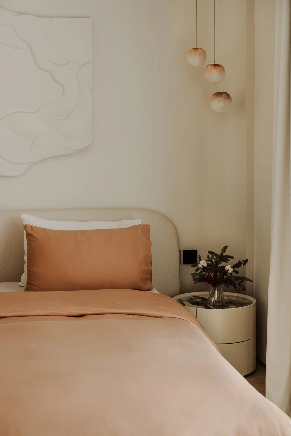 Luxury bedroom with terracotta satin bedding, a bouclé curved headboard, a sculptural white relief wall artwork, and amber glass cluster pendant lights, designed by Limestone in Dubai