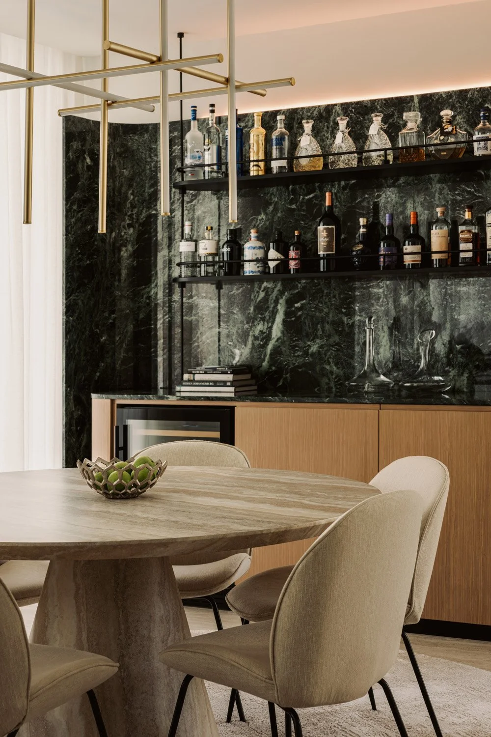 Luxury dining room featuring a travertine round table, upholstered chairs, green marble bar wall, and sculptural brass chandelier by Limestone