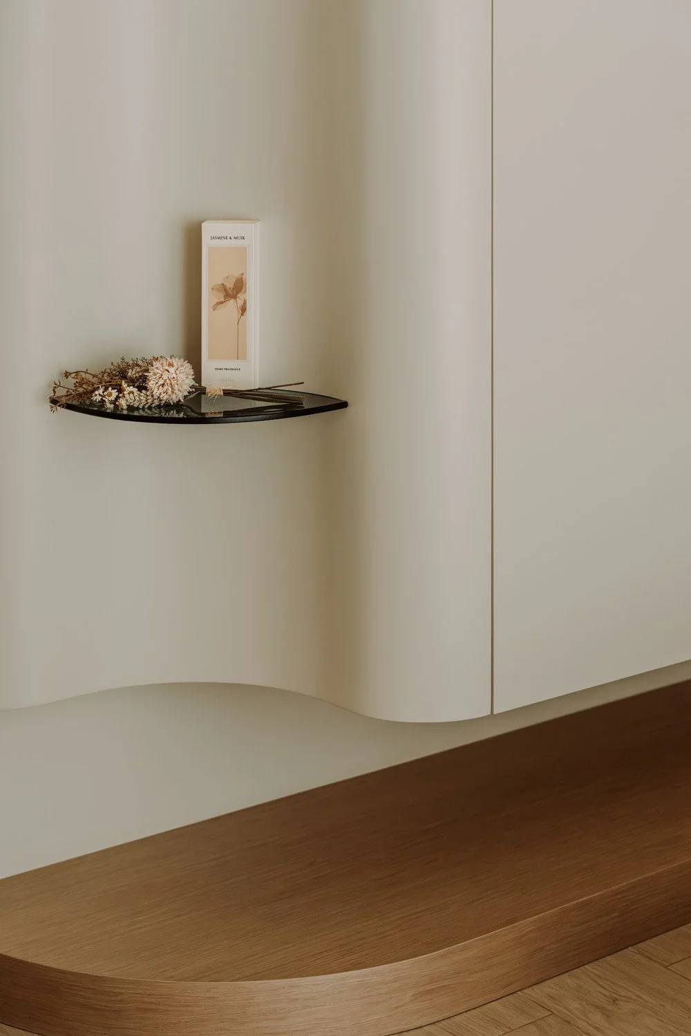 Minimalist bedroom detail featuring a floating smoked glass shelf with dried floral arrangement and fragrance diffuser, set against curved white plaster joinery and an oak platform base, designed by Limestone