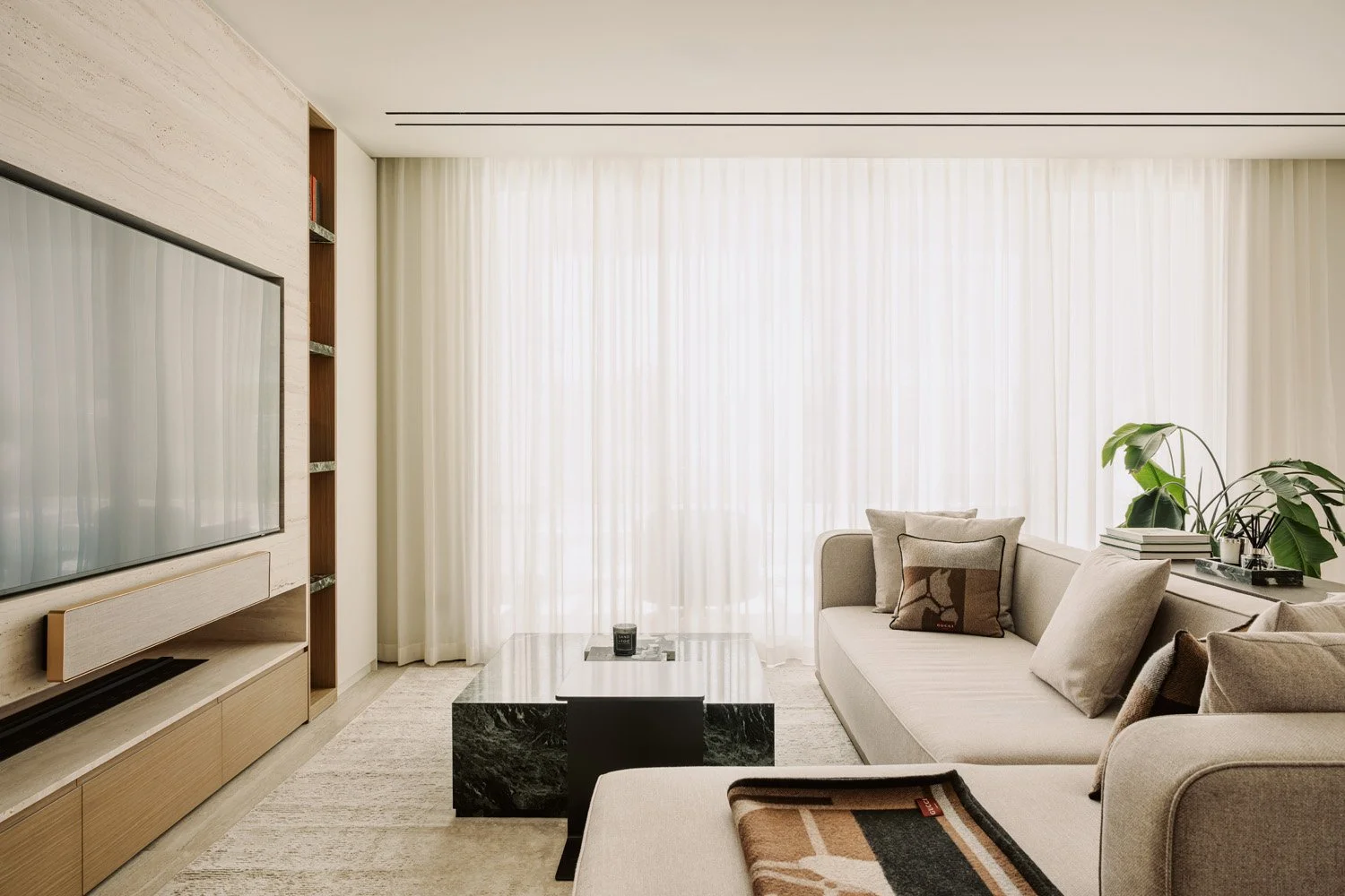 Luxury living room with a travertine-clad media wall, built-in timber shelving, sectional sofa, and floor-to-ceiling sheer curtains by Limestone