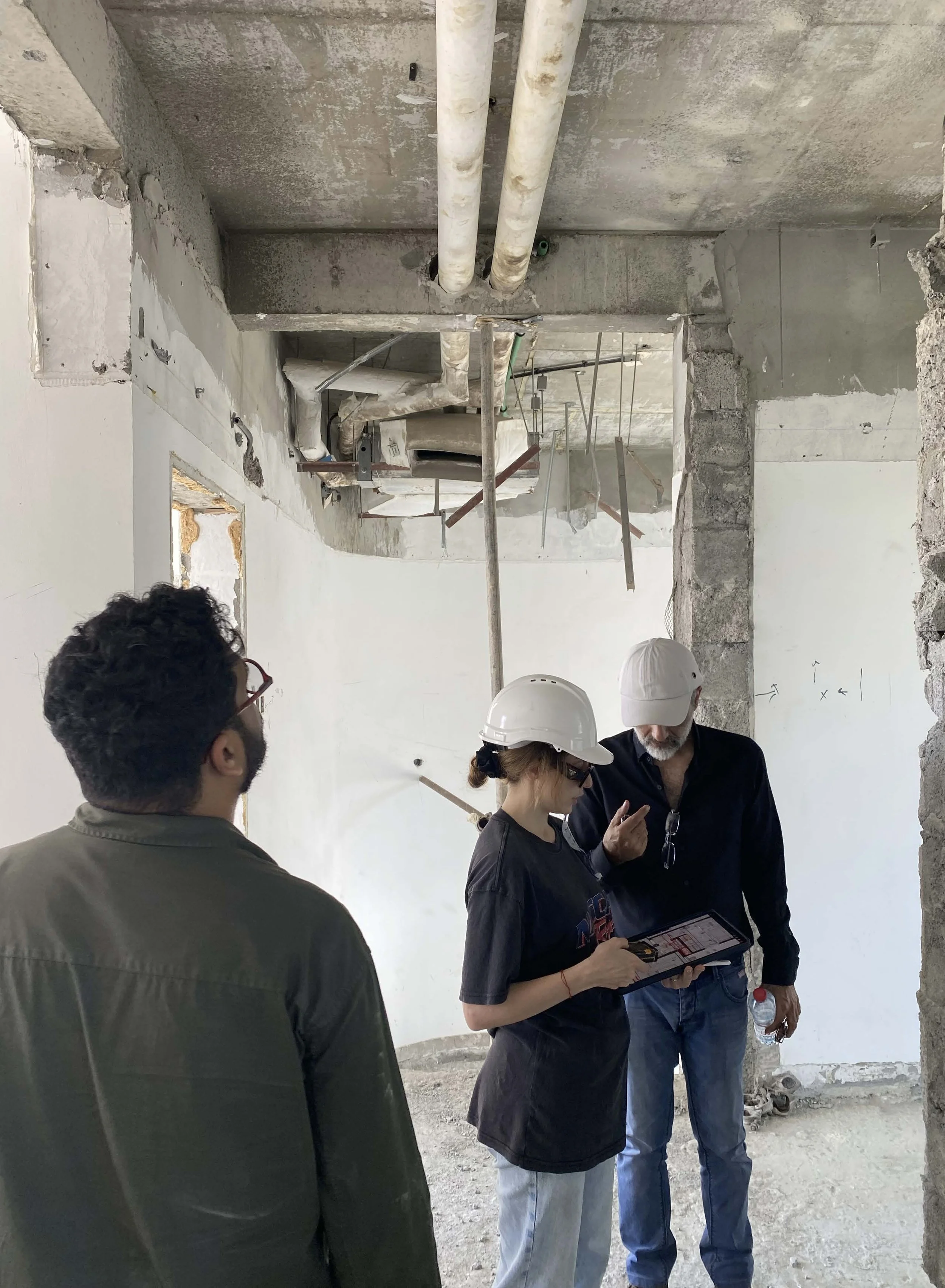 Three construction workers, two men and one woman, examining plans inside an unfinished building with exposed pipes, concrete walls, and ceiling.