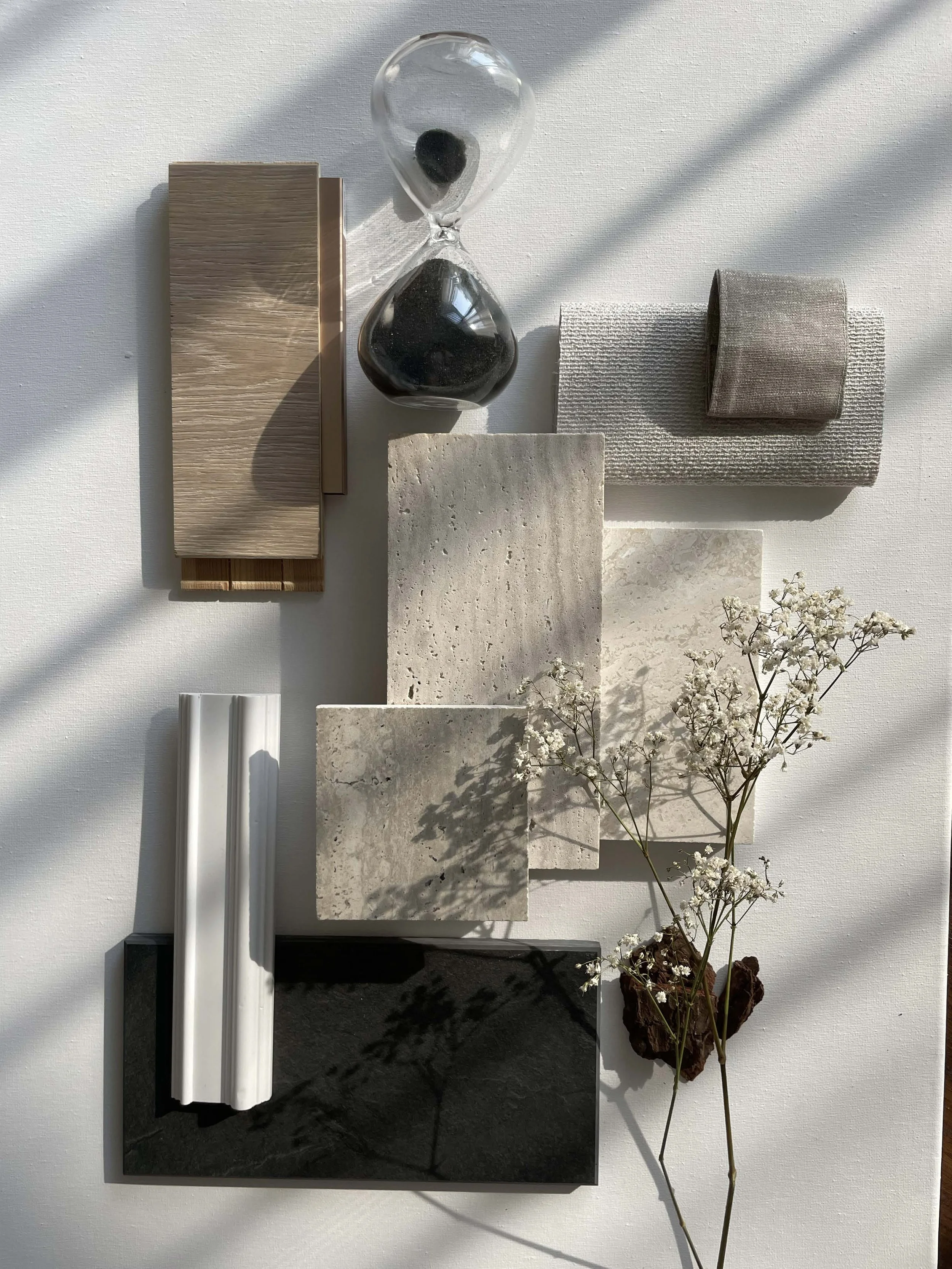 Arrangement of various textured swatches, a glass hourglass filled with black sand, a flower with small white blossoms, and shadows on a white background.