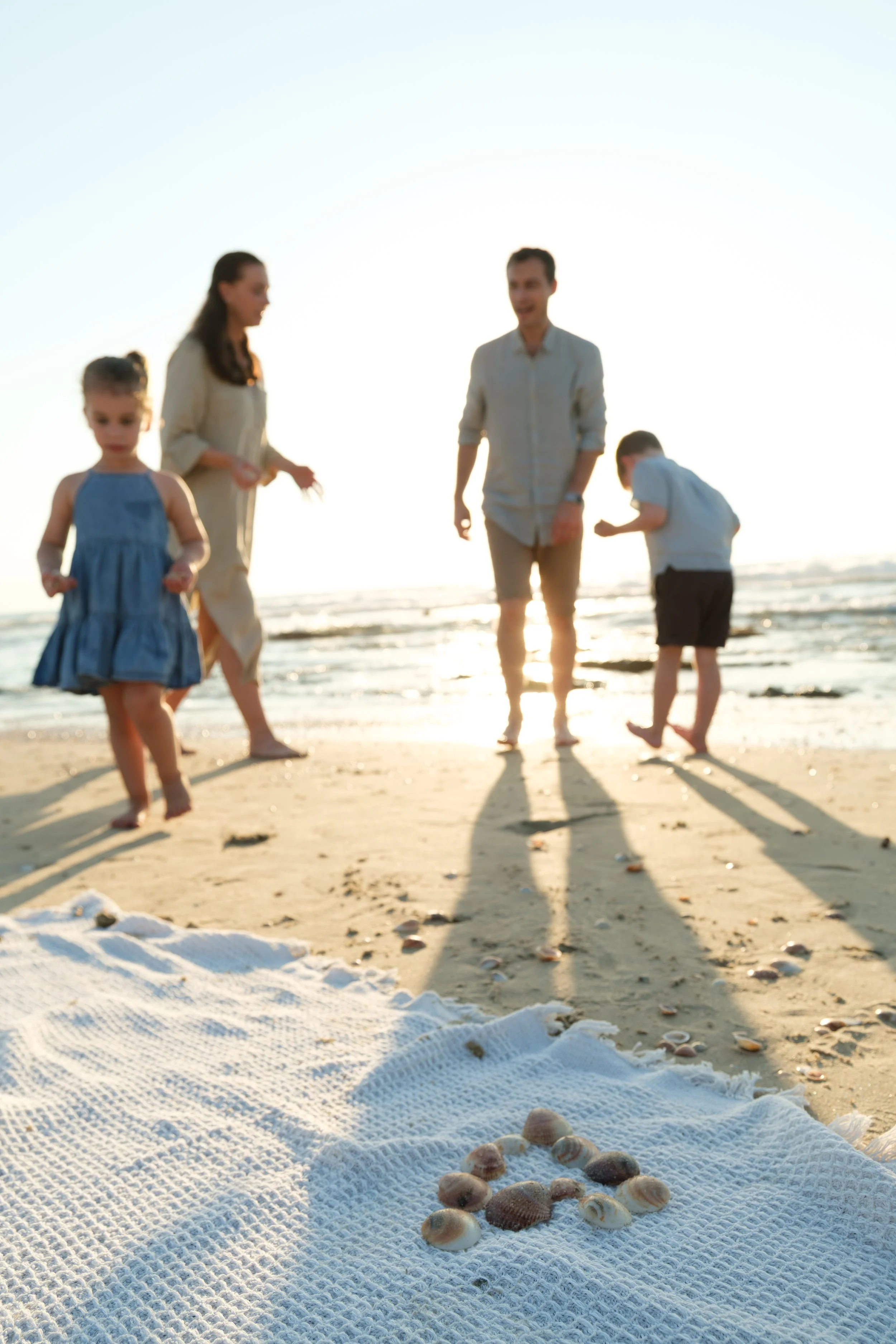 Scene of four people walking on the beach near the water at sunset with shadows cast on the sand by the sunlight. In the foreground, there is a white towel with seashells on it.