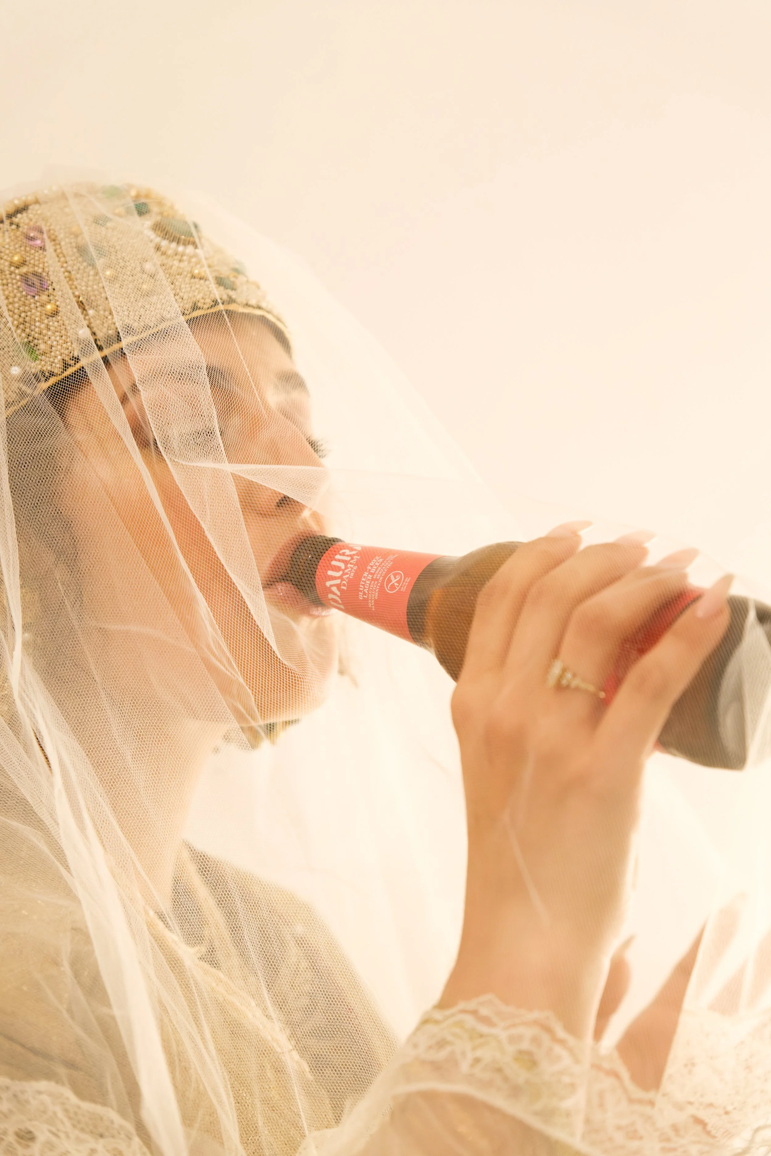 A woman dressed in a wedding gown and veil is holding a bottle of champagne, taking a sip.