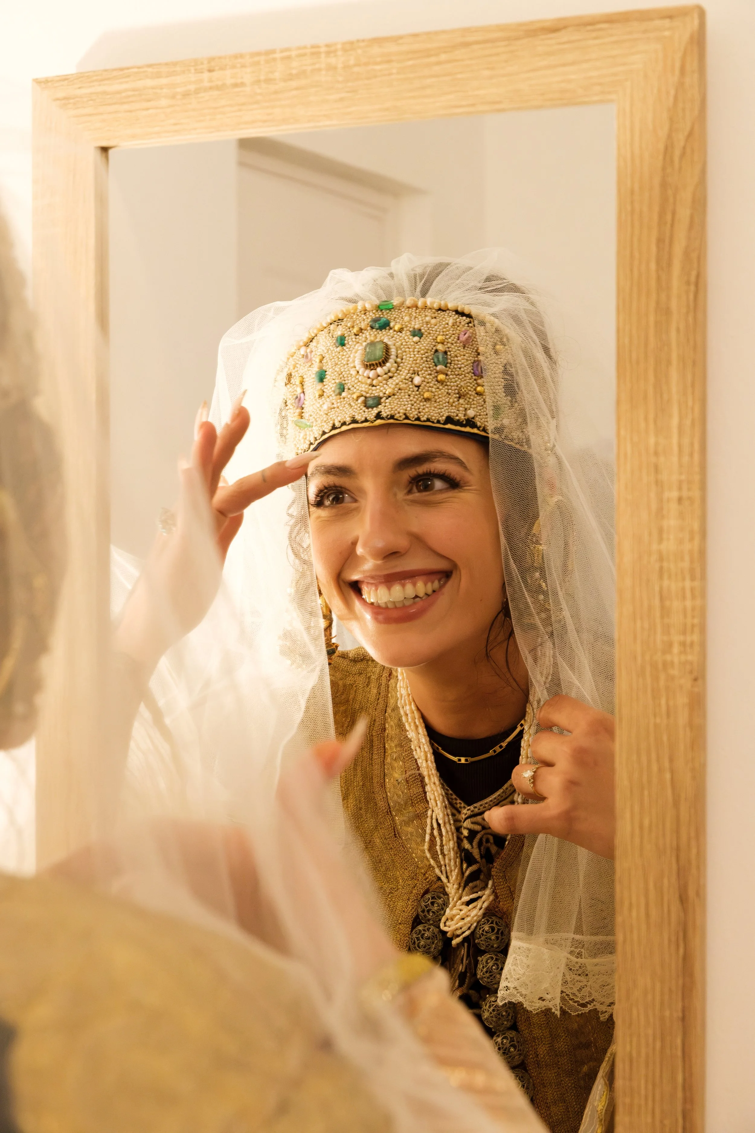 A woman looking at herself in a mirror, wearing a beaded headdress with pearls and gemstones, a veil, and traditional clothing, smiling.