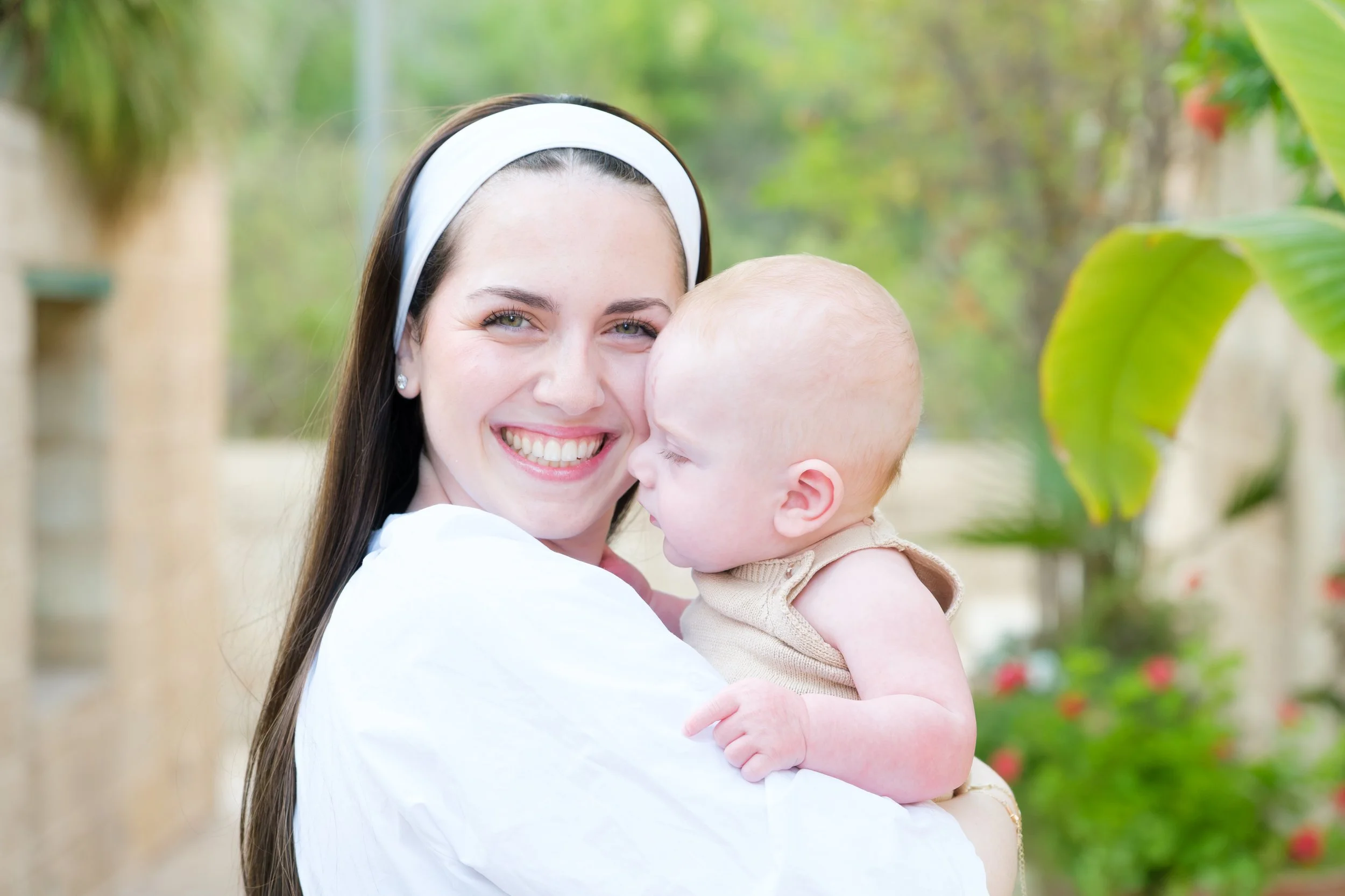 A woman with long brown hair, wearing a white headband and white shirt, smiling and holding a baby with light skin and light-colored hair, outdoors with green foliage in the background.