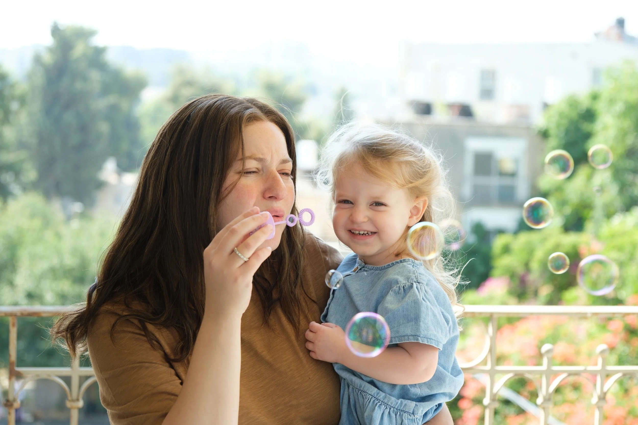 A woman with long brown hair and a young girl with blonde hair blowing bubbles outdoors on a sunny day, with greenery and buildings in the background.