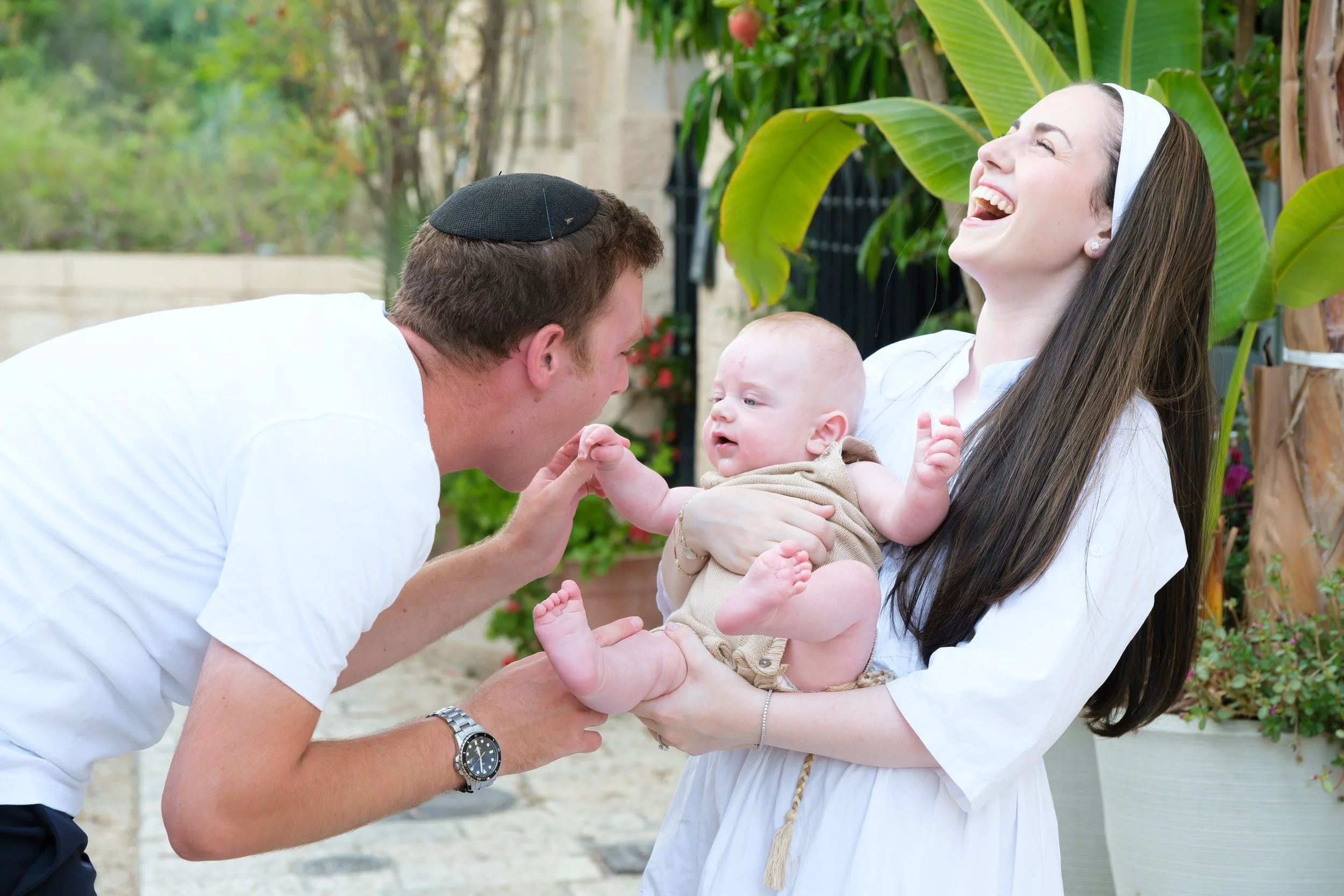 A woman with long brown hair, wearing a white headband, is holding a baby in her arms. A man wearing a black kippah and a white shirt is reaching out and holding the baby's hand. They are outdoors, surrounded by green plants and trees, and are smiling and laughing together.
