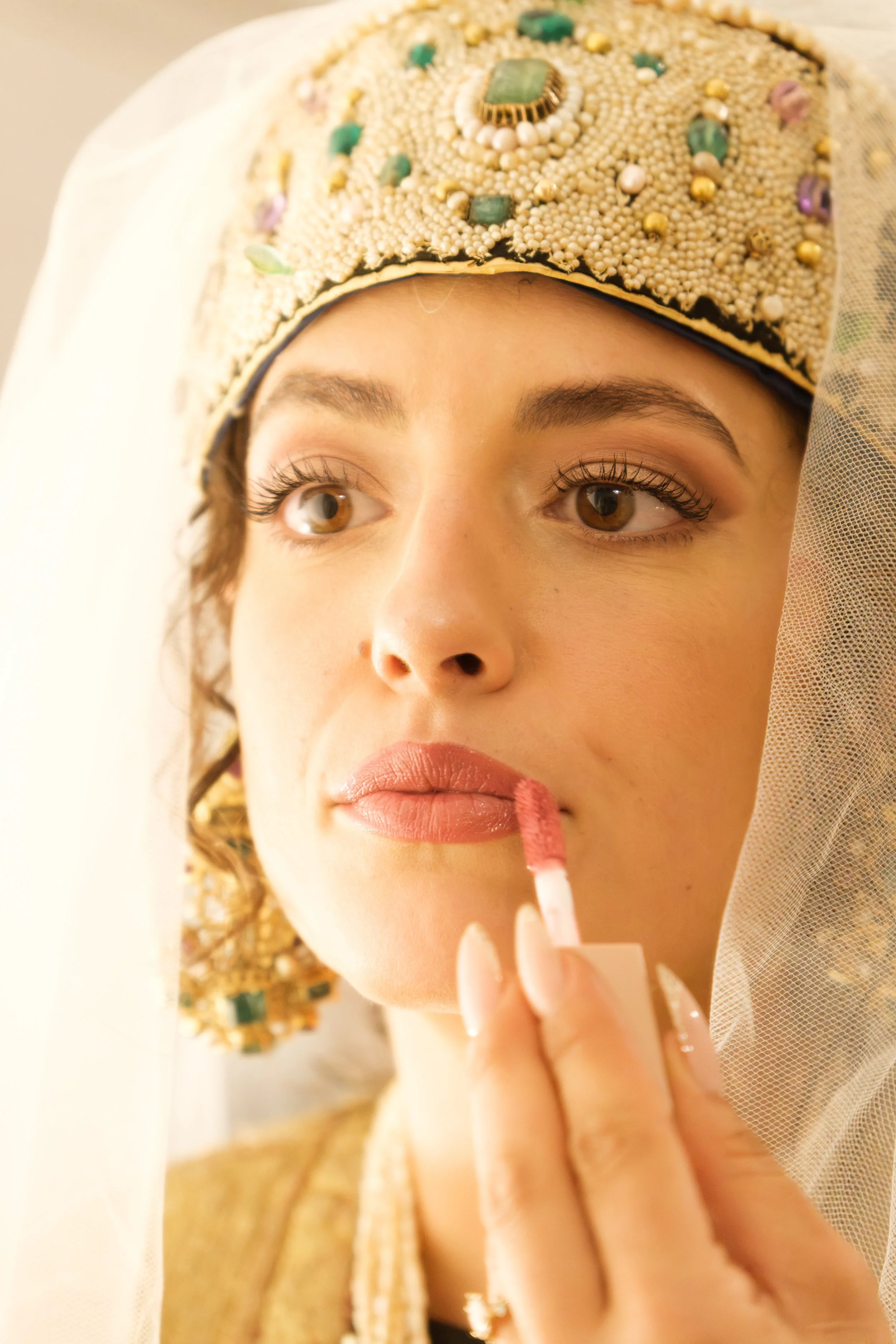 A woman with detailed, ornate jewelry and a decorative headpiece, applying makeup using a lip gloss applicator.