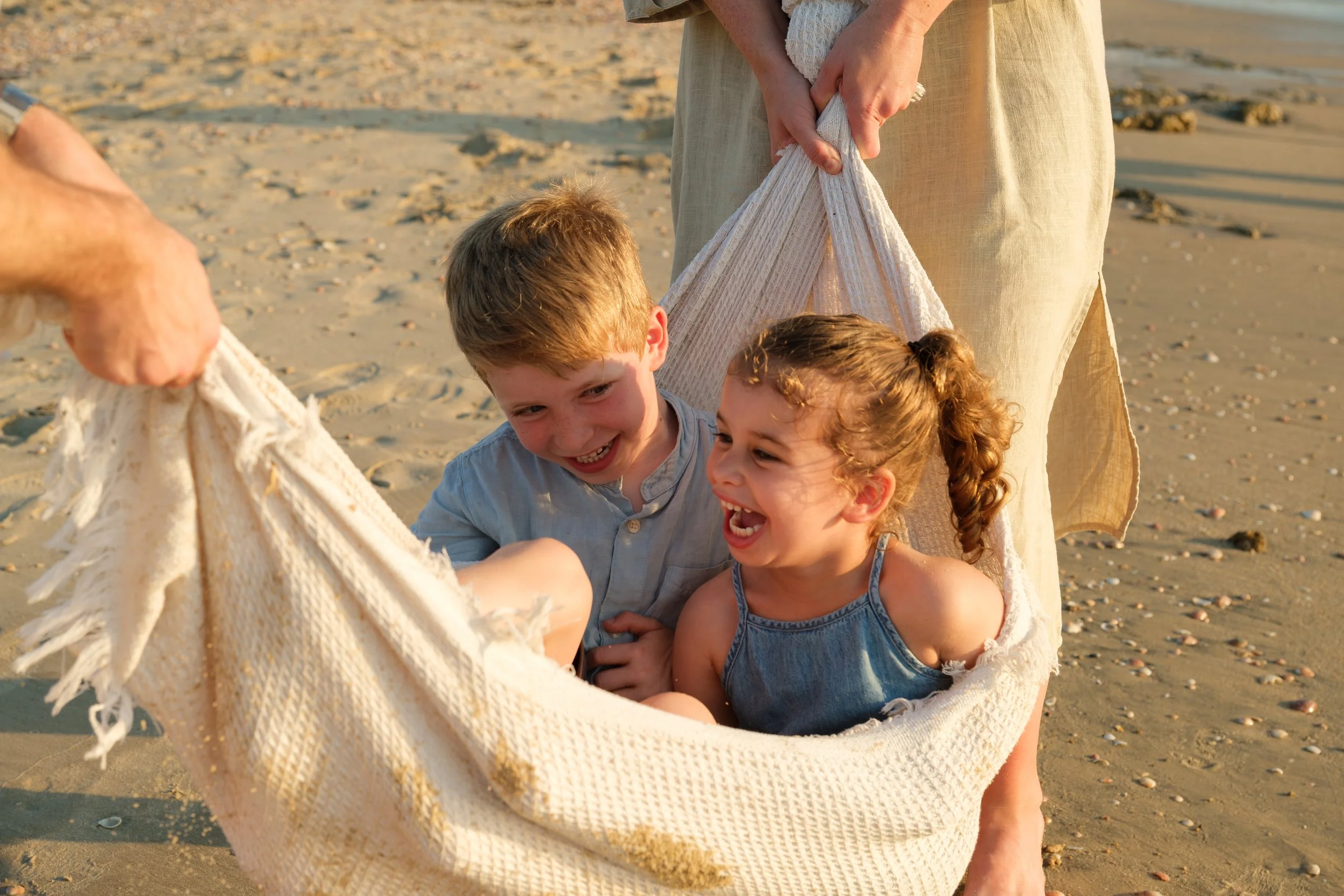 Two children, a boy and a girl, smiling and laughing as they hold onto an adult's hand and play in a hammock on a sandy beach during sunset.