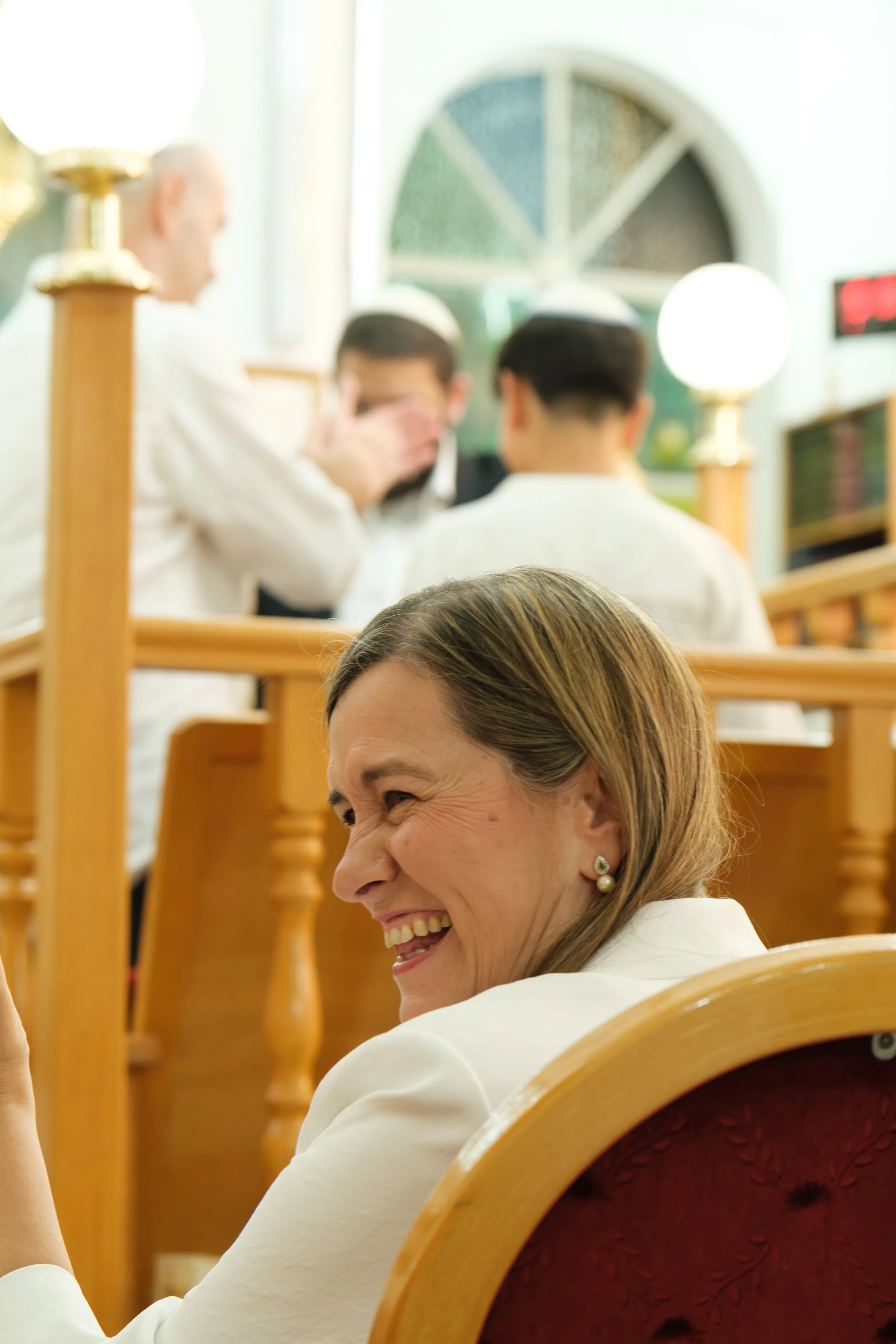 A woman with blonde hair smiling and laughing, seated in a wooden chair with a red velvet back, in a room with other people in white attire, possibly a religious or communal gathering.
