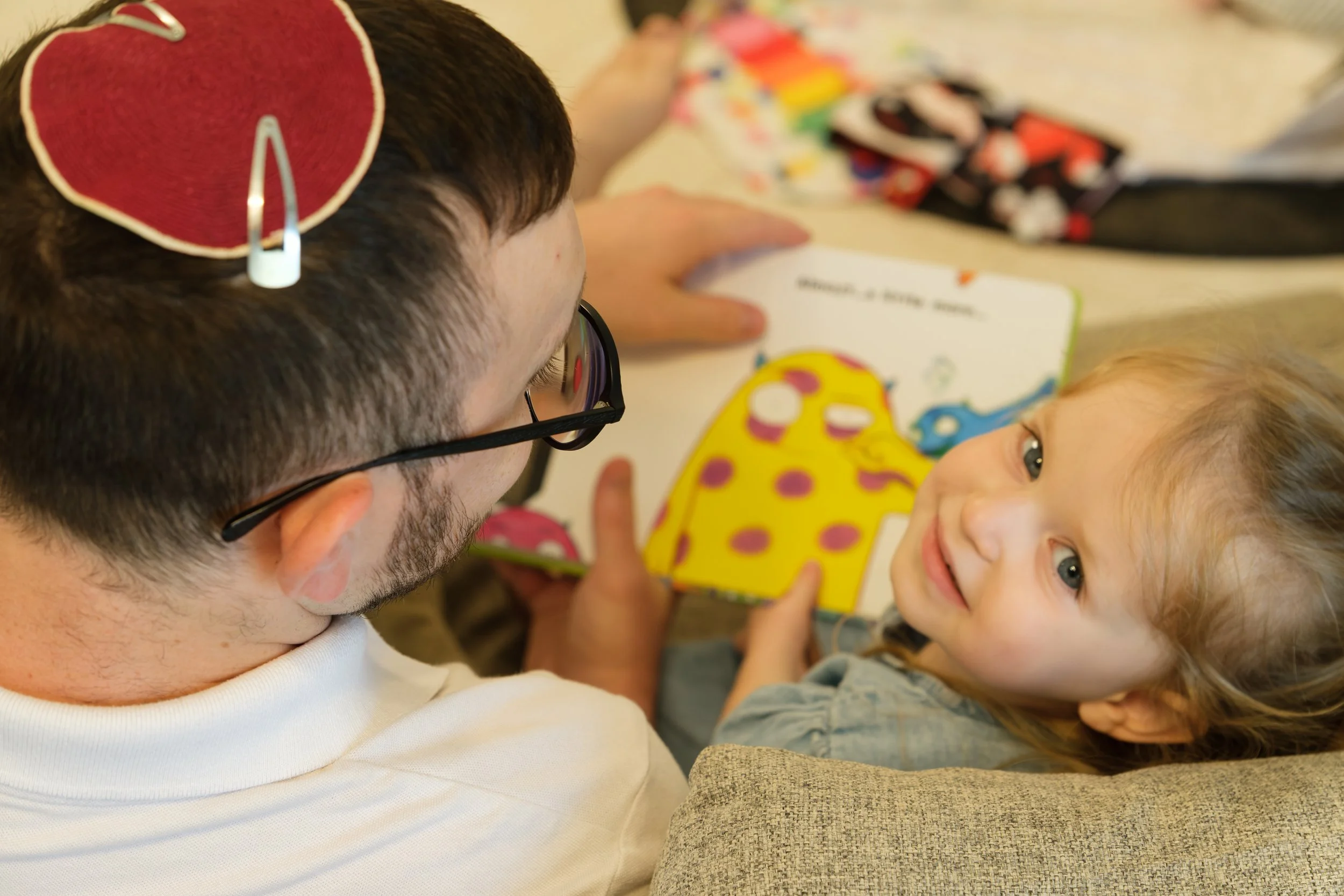A man with glasses and a beard reading a children's book to a young girl with light hair and bright eyes, sitting on a beige couch in a cozy setting with a colorful blanket nearby.