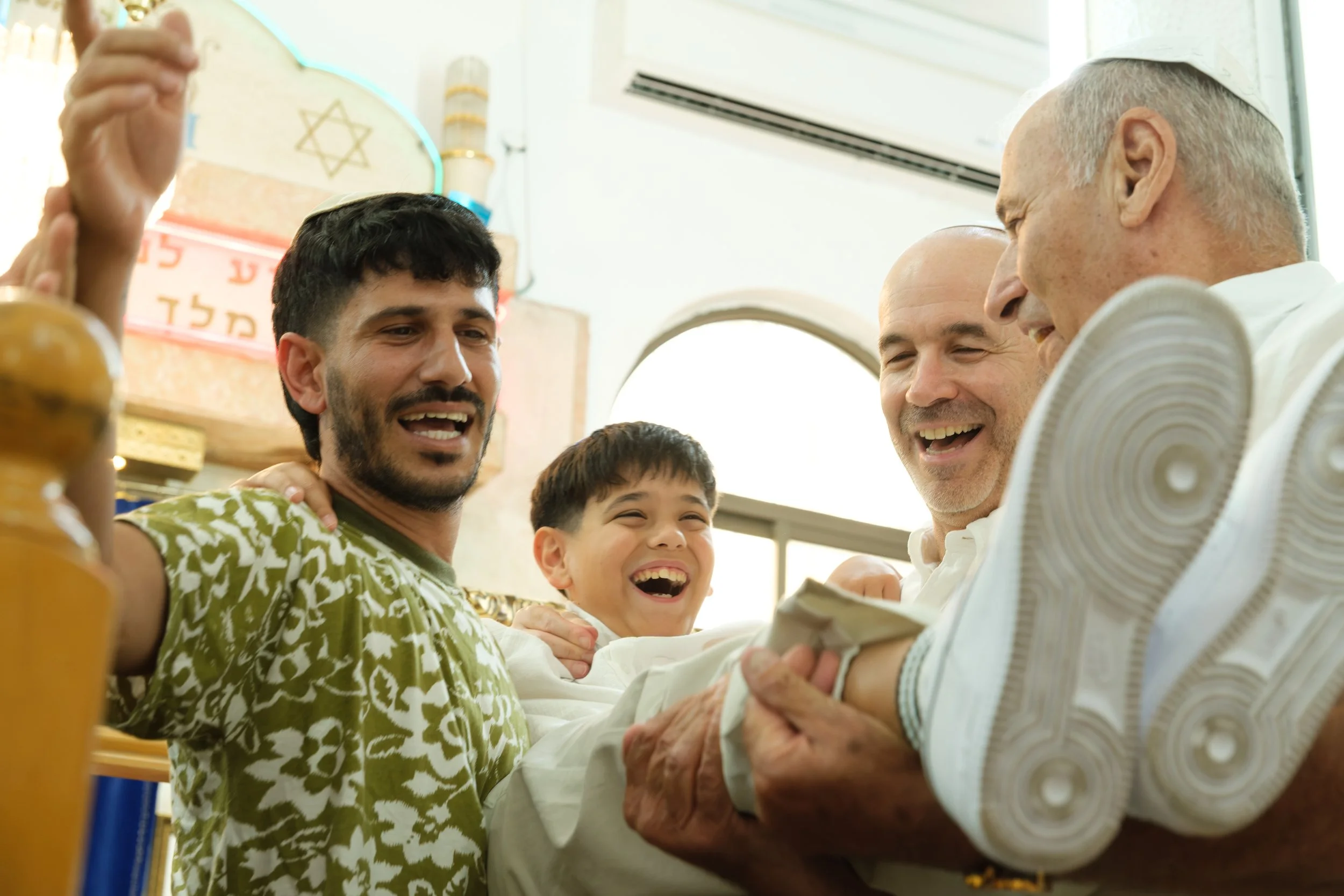 A diverse group of people, including a young man, two older men, and a young boy, smiling and gathered around, possibly in a religious or cultural setting, with some individuals wearing traditional Jewish attire.