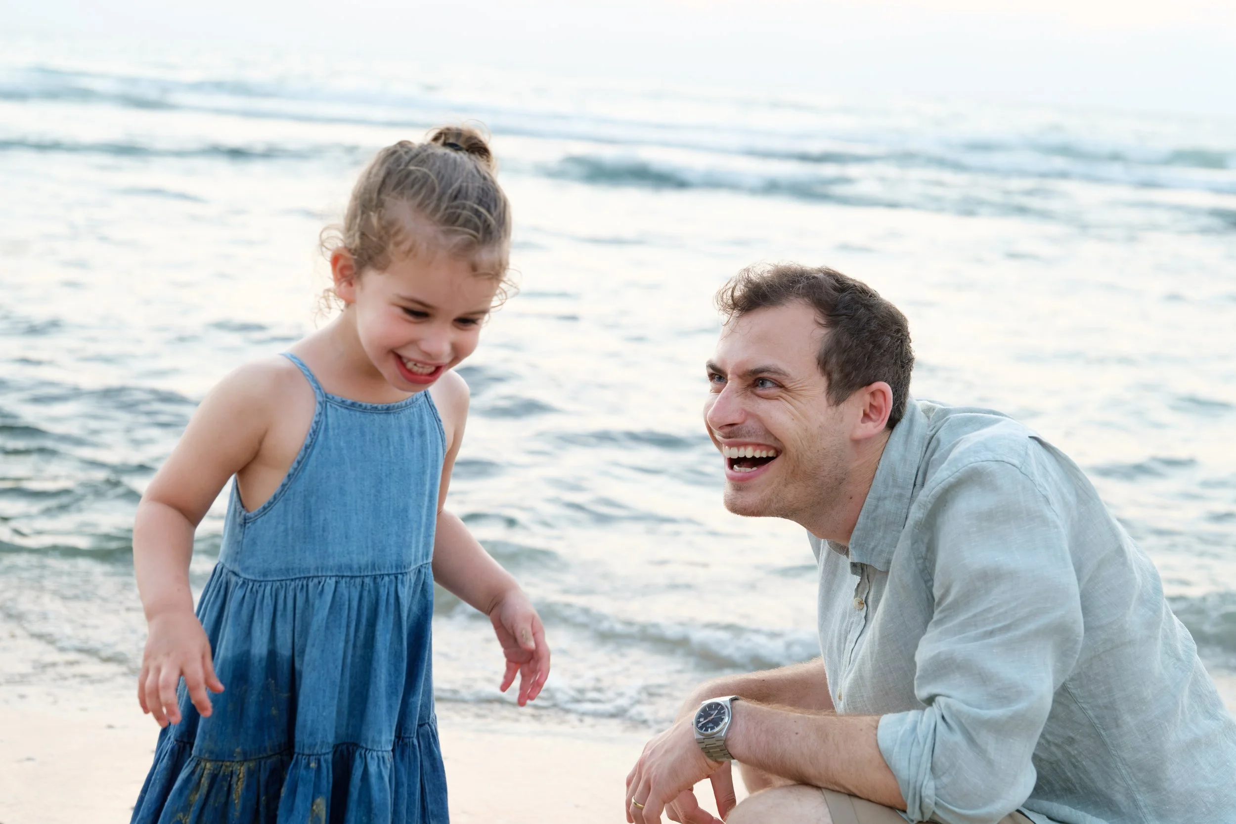 A young girl in a blue dress smiling and looking down at the beach, while an adult man, possibly her father, crouches nearby smiling at her with the ocean in the background.